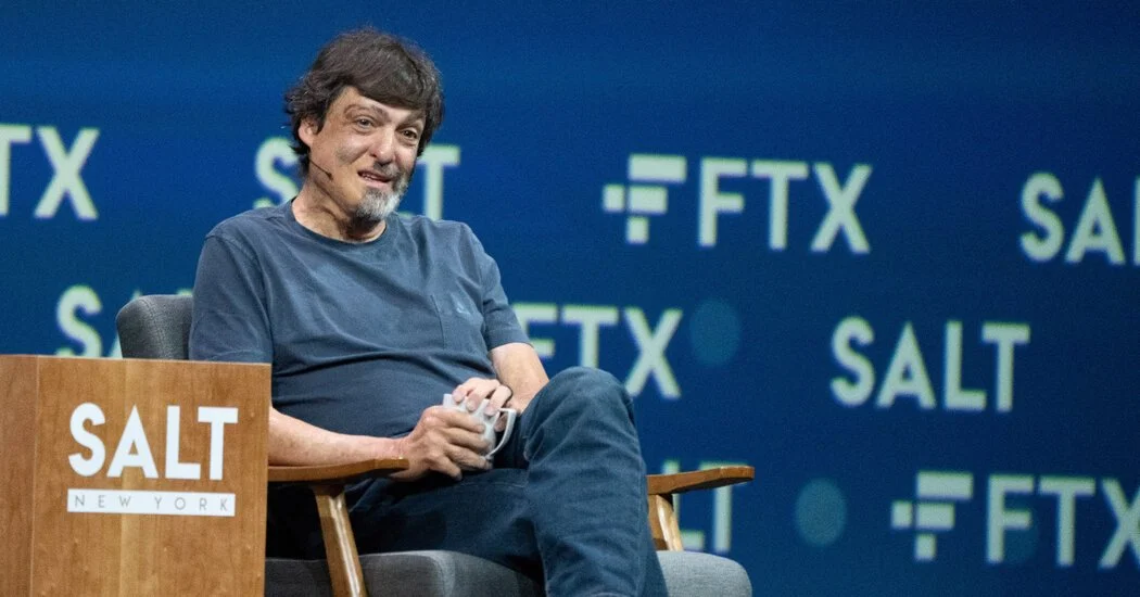 A man with dark hair and a beard sitting in a chair at a conference, holding a mug, with a backdrop displaying the words 'SALT' and 'FTX' in blue and green text.