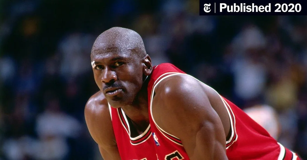 Close-up of a basketball player in a red jersey, looking intense, after a game, with a blurry crowd in the background.