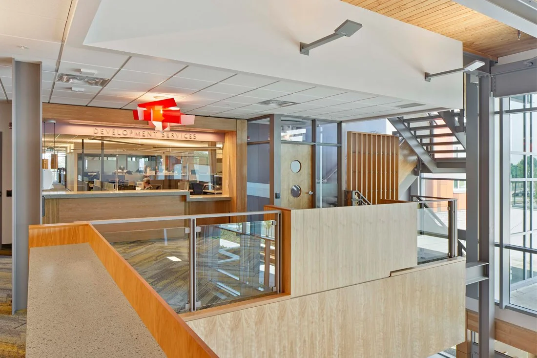 Interior view of an office building with a reception area labeled 'Development Services,' wooden and glass railing, staircases, and large windows letting in natural light.