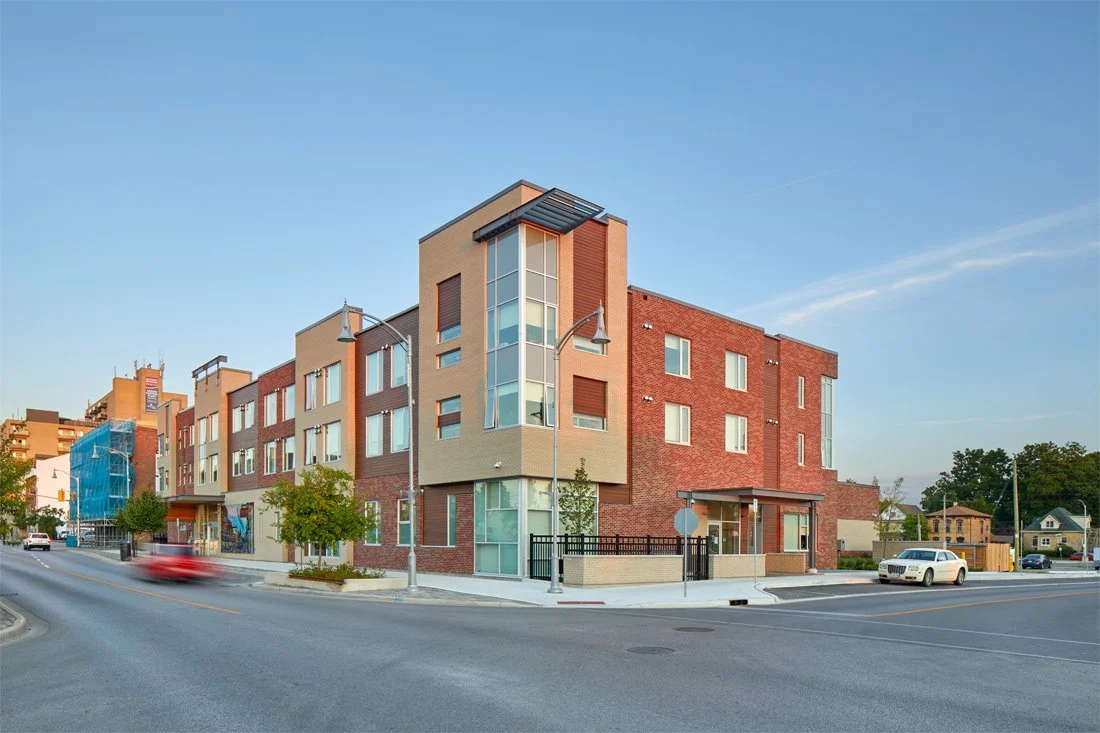 Multi-story modern residential building on a city street with trees and cars parked nearby under a clear blue sky.