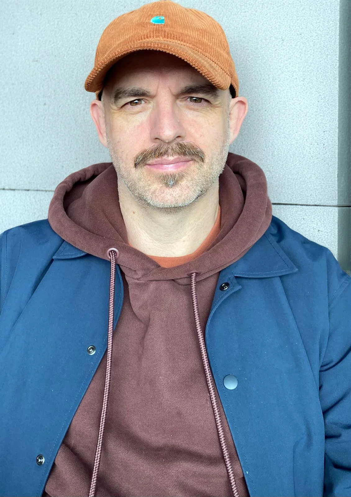 A man wearing an orange cap, brown hoodie, and blue jacket taking a selfie outdoors against a light gray wall.