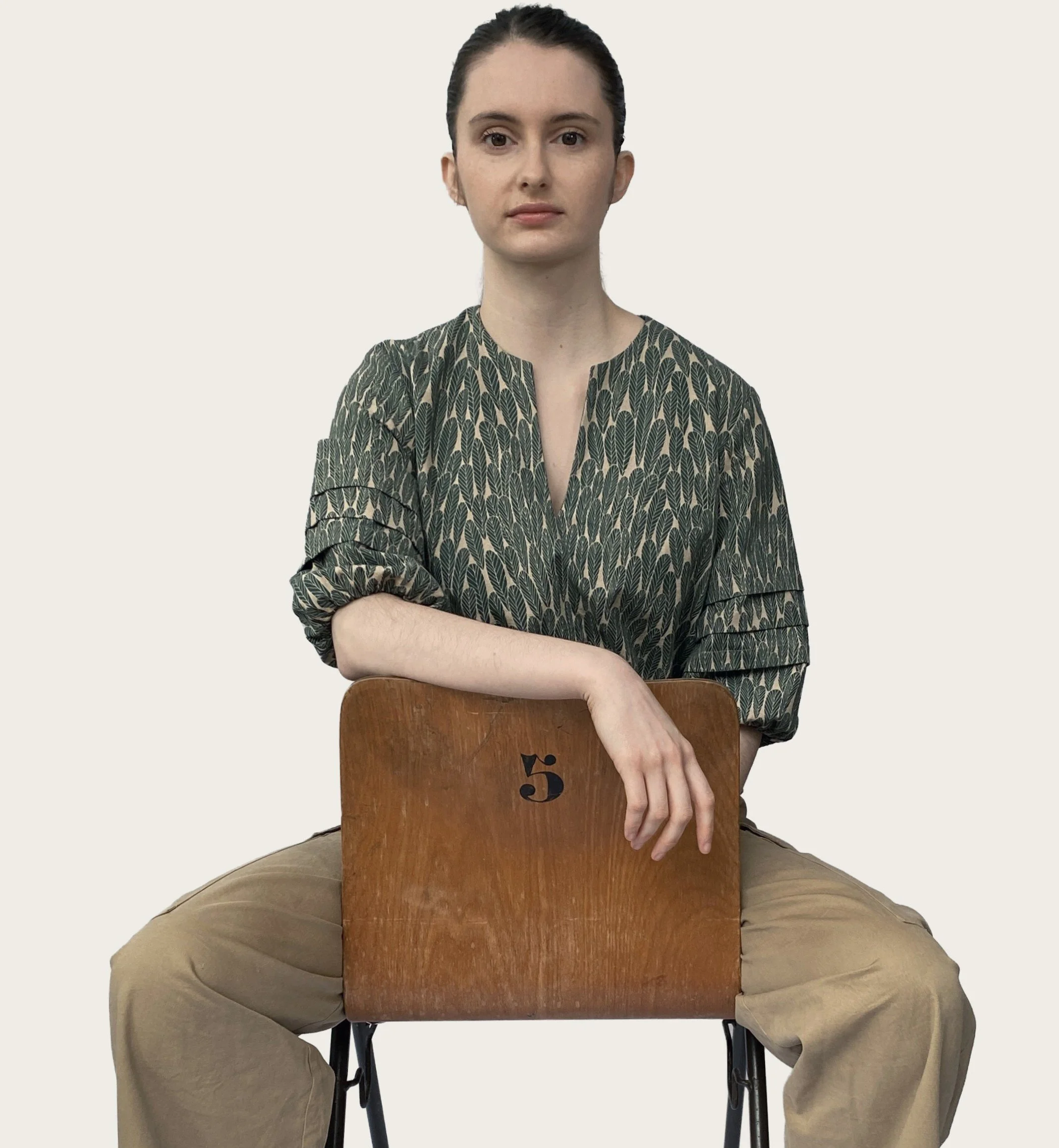A woman sitting on a chair against a plain white background, her left arm resting on the back of the chair which has the number 5 written on it.  Wearing the Claudia Top sewn by SewClothing using the Claudia Top Sewing Pattern.