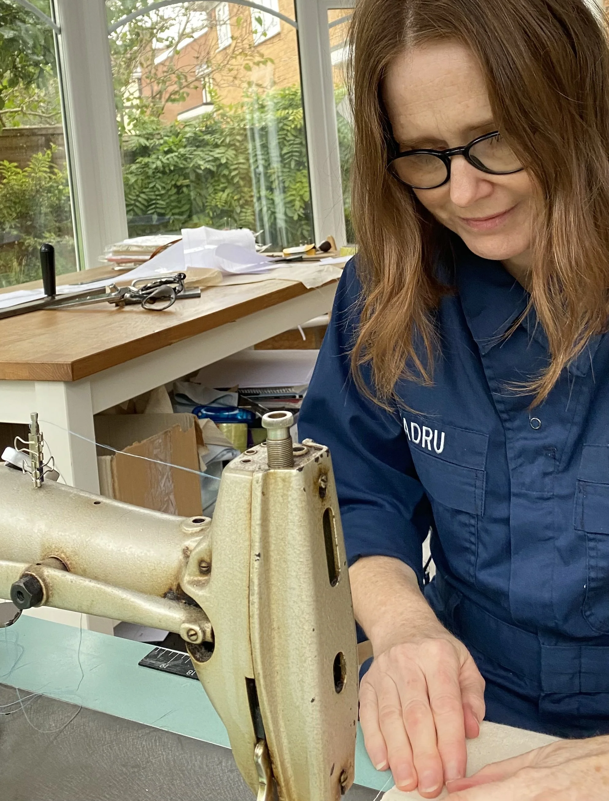 A woman wearing glasses and a blue uniform working with a sewing machine in a bright room with large windows and green foliage outside.