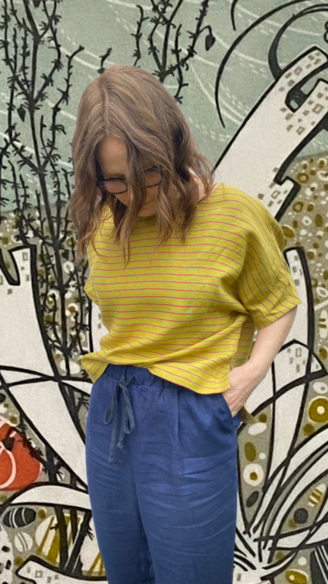 A woman with wavy brown hair and glasses wearing a yellow and red striped top made from sewclothing elena top pattern and blue drawstring pants, made from sewclothing elena trouser pattern standing in front of an illustrated background.