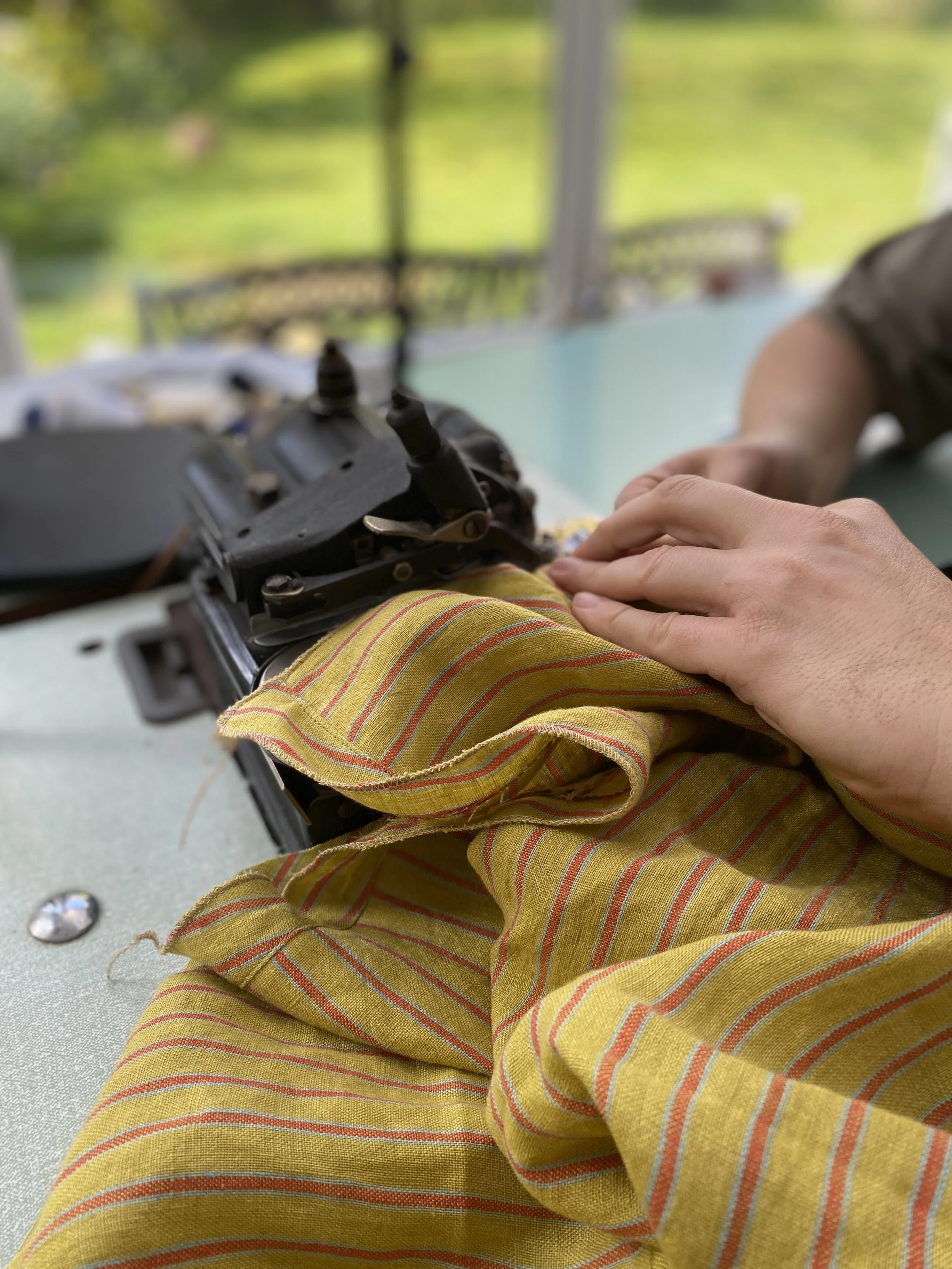 A person sewing yellow and orange striped fabric making the sewclothing elena asymmetric top pattern with a blurred green garden background.
