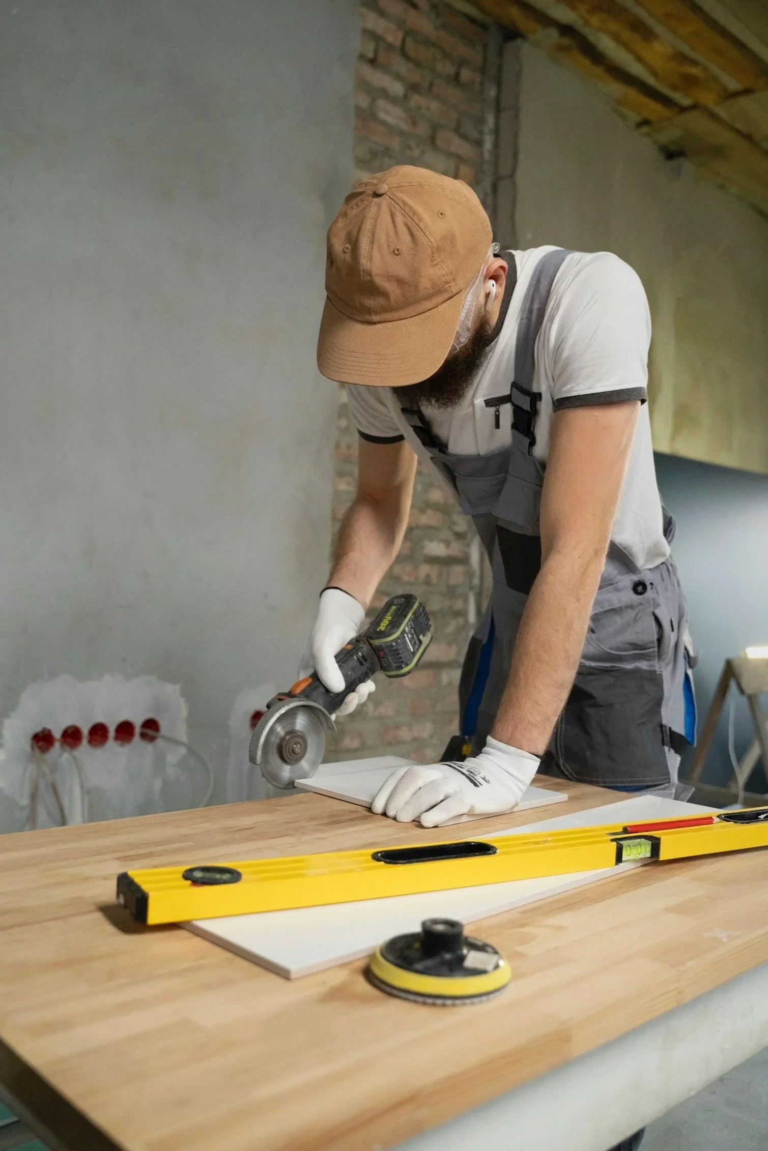 A man wearing a brown cap, gray overalls, and white gloves is using a cordless circular saw to cut a white board on a wooden workbench in a construction or renovation site.