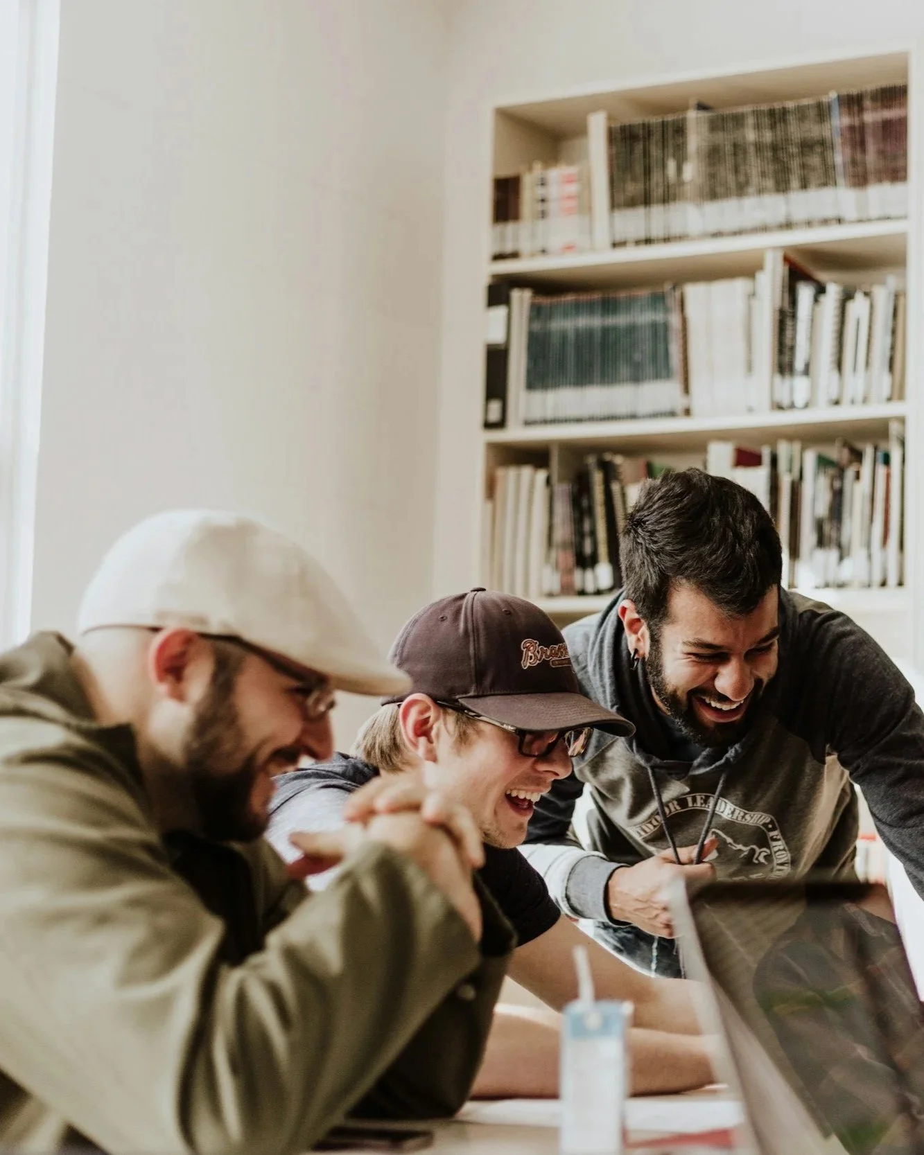 Three men laughing and looking at a laptop in a room with a white bookshelf filled with books in the background.