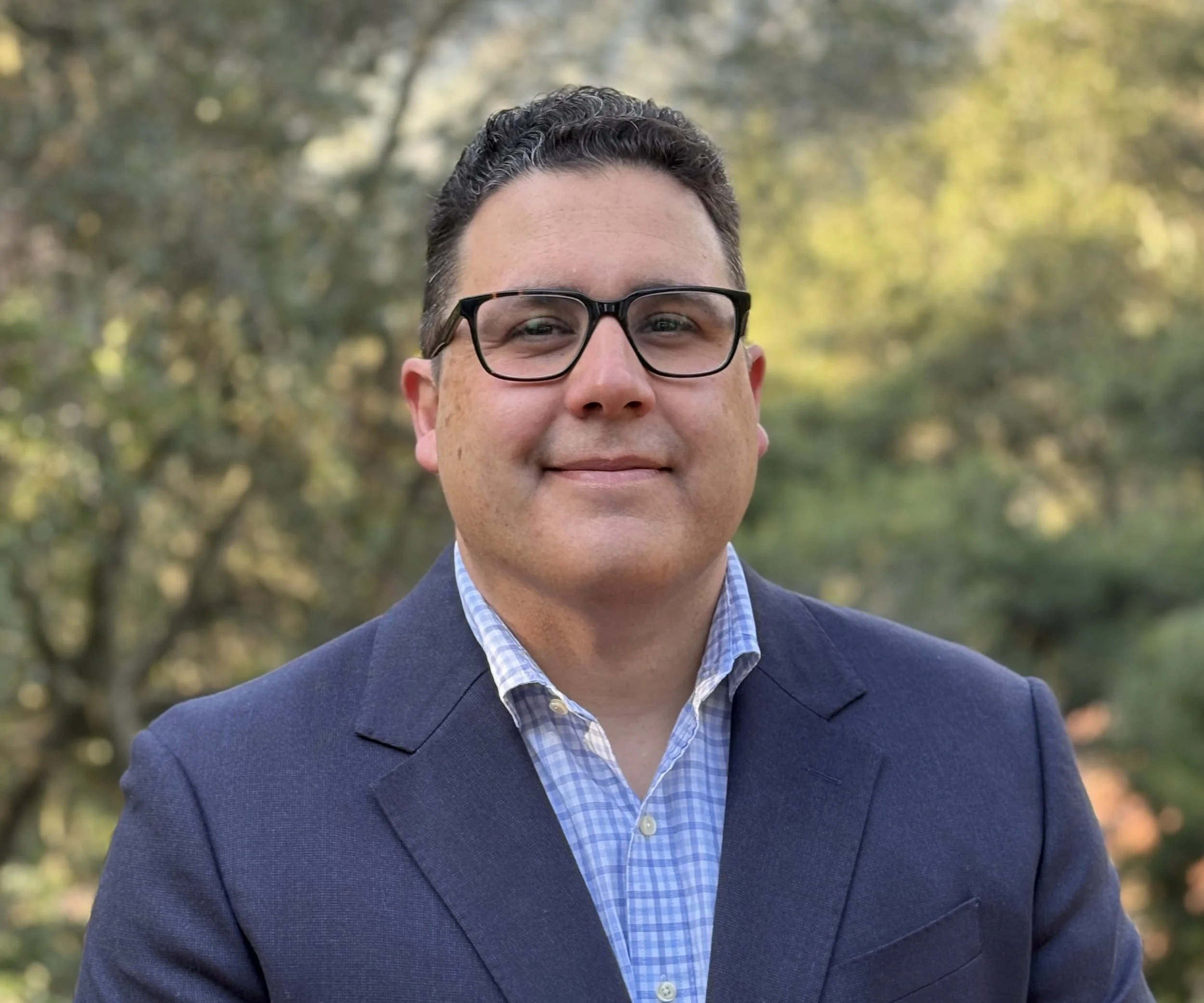 Portrait of a man wearing a blue suit and glasses outdoors in a green park during daytime.