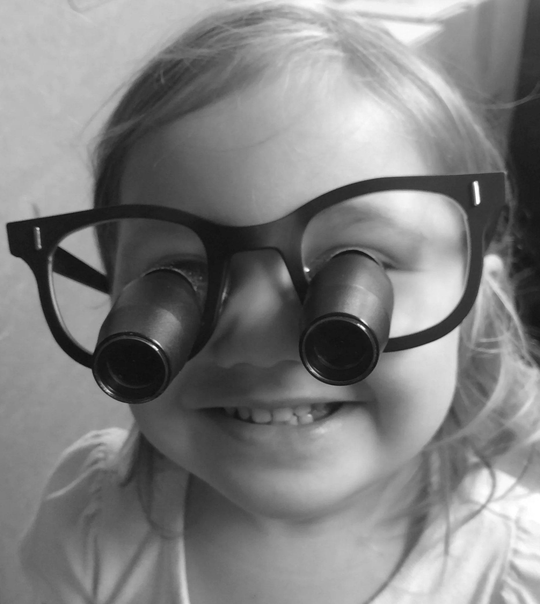 A young girl wearing glasses with two magnifying lenses attached, smiling and looking through the lenses.