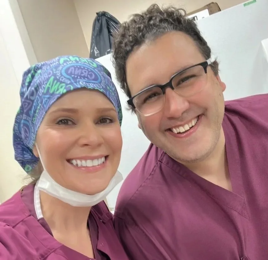Two healthcare professionals, a woman with a colorful scrub cap and a man with glasses, smiling in a medical setting.