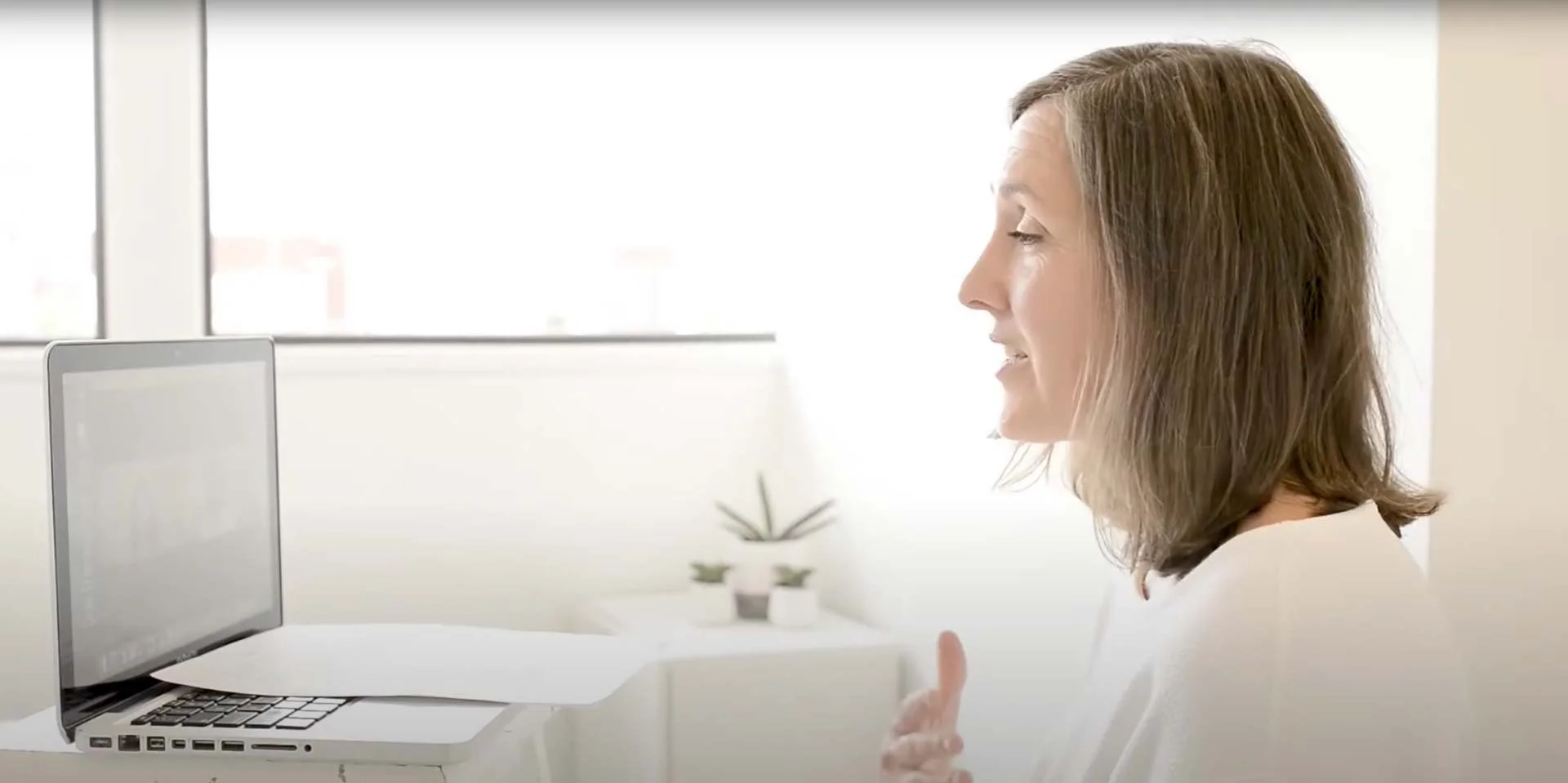 Side view of a woman with brown hair smiling and gesturing in front of a laptop on a white desk, in a bright room with large windows and a small potted plant in the background.