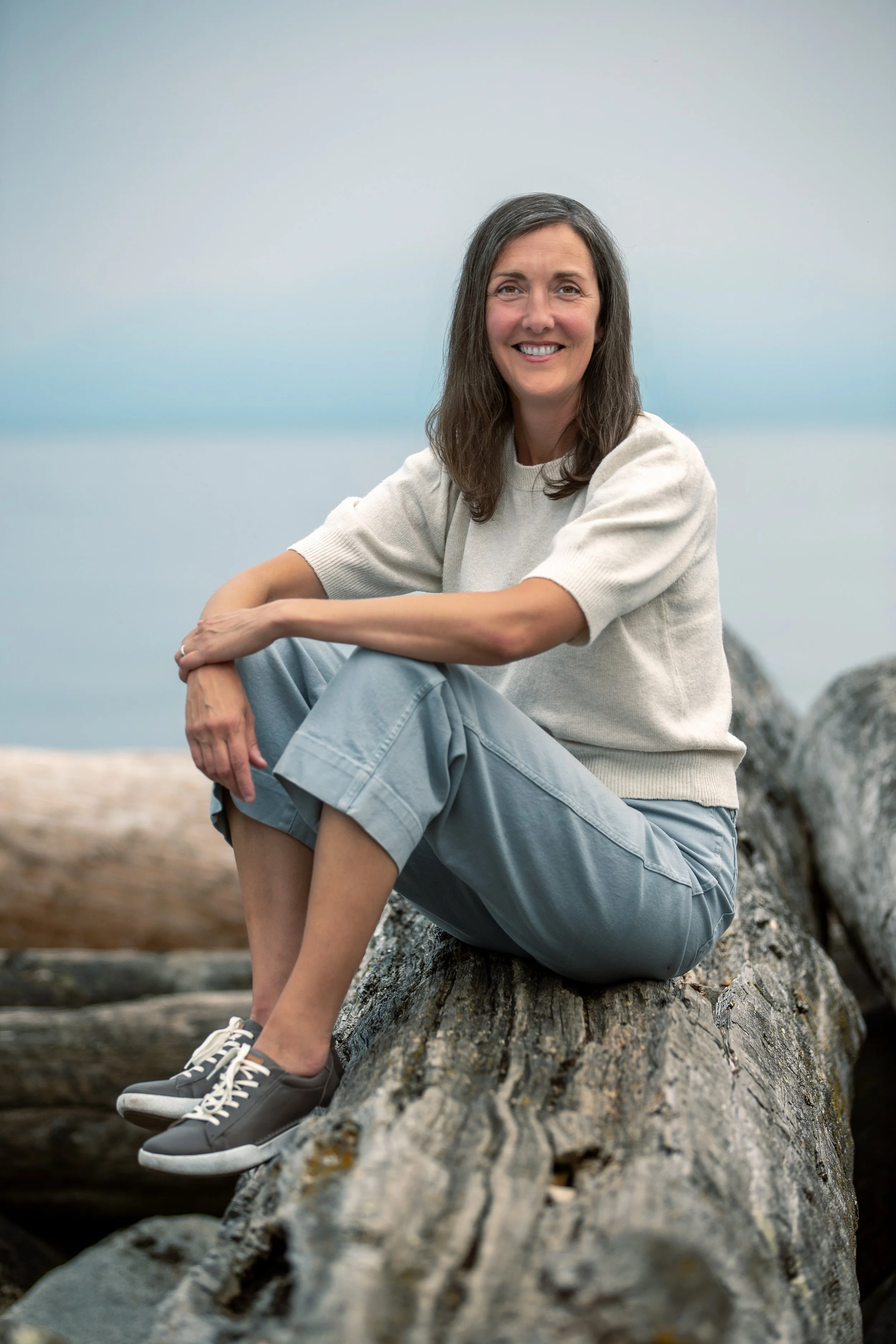 A woman with long dark hair, sitting on a large log near the water, smiling at the camera. She is wearing a light-colored sweater, gray pants, and gray sneakers.