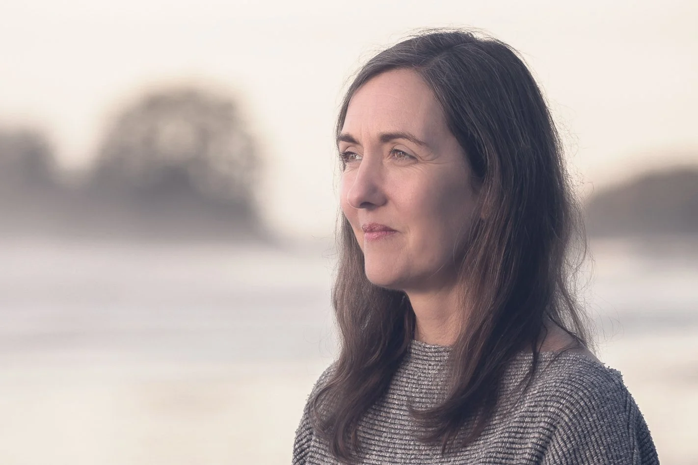 A woman with long dark hair looking into the distance outdoors with blurred trees and sky in the background.