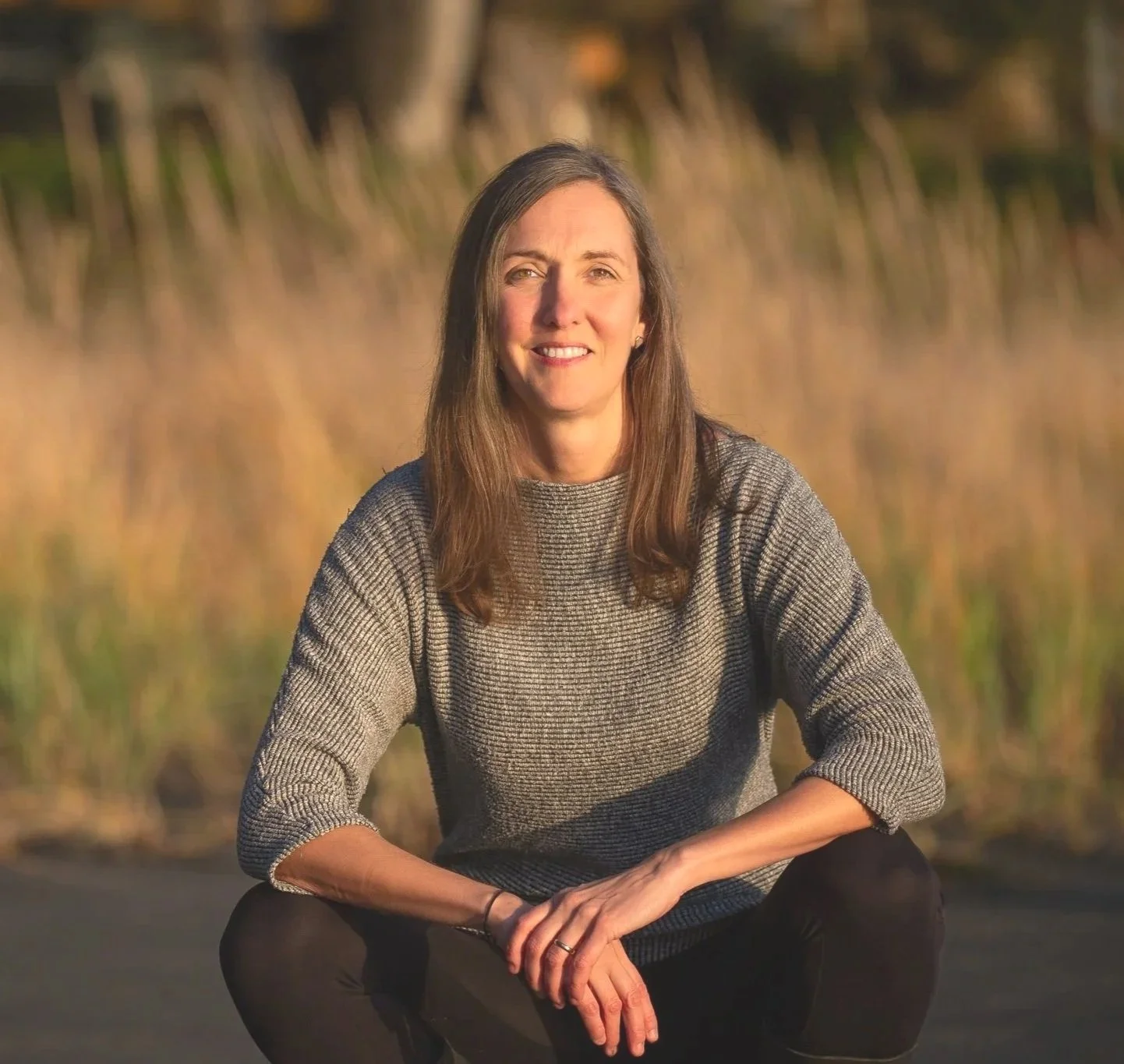 A woman with long brown hair, wearing a gray knit sweater, sitting outdoors on a road with tall grass and trees in the background during golden hour.
