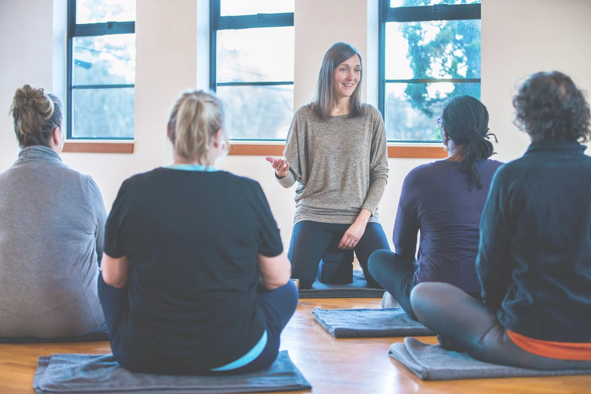 A woman leading a yoga or meditation class, sitting on a mat in front of five women who are seated on similar mats, in a room with large windows and wooden floors.