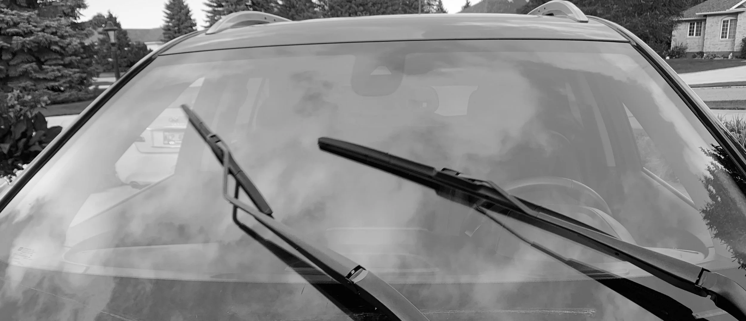 Black and white photo of a car's windshield with its wipers in the resting position, with houses and trees visible in the background.
