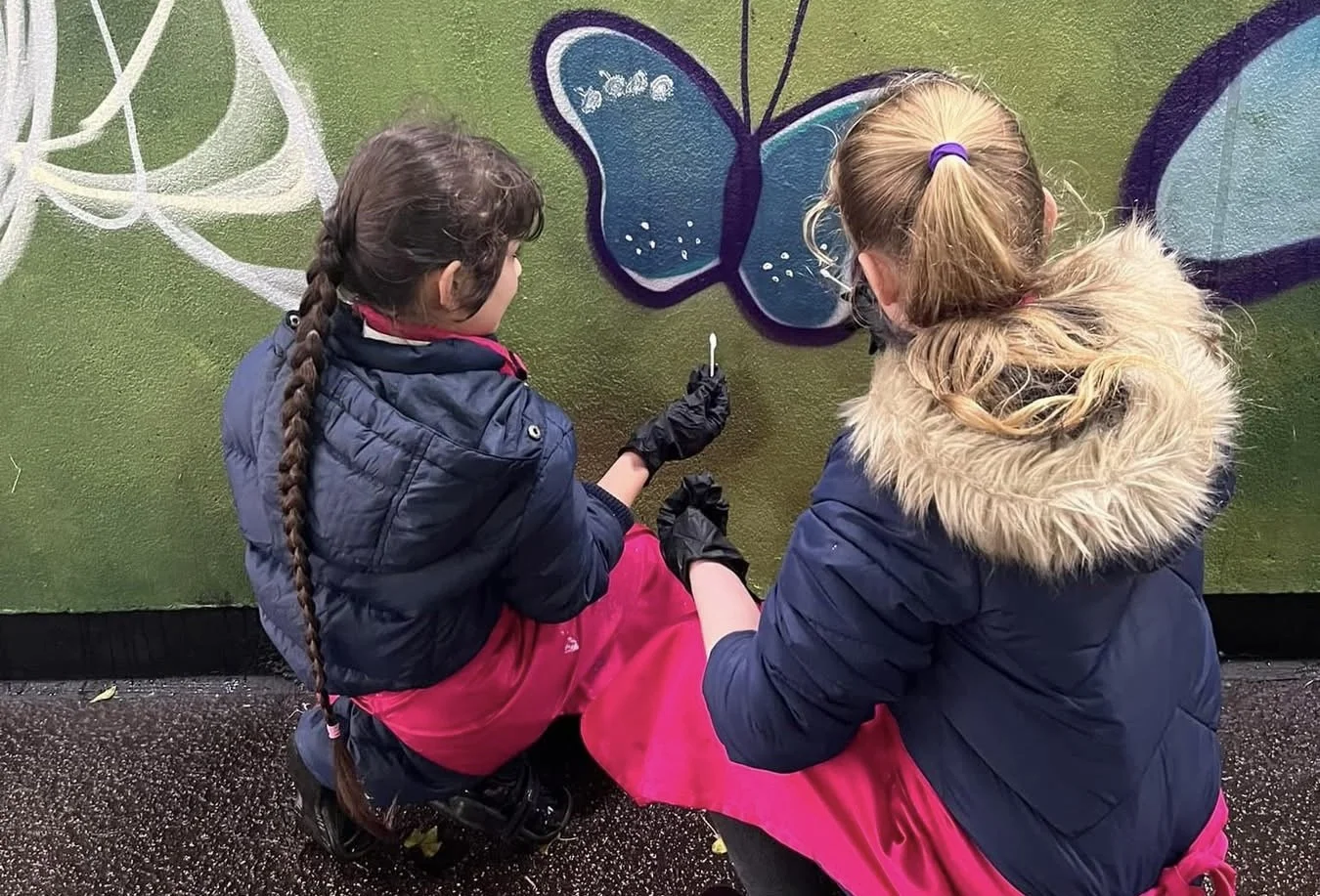Two girls sitting on the ground, painting a blue butterfly on a wall. One girl has a long braid and is holding a paintbrush, the other has a ponytail with a purple hair tie. They are dressed in winter jackets and gloves.