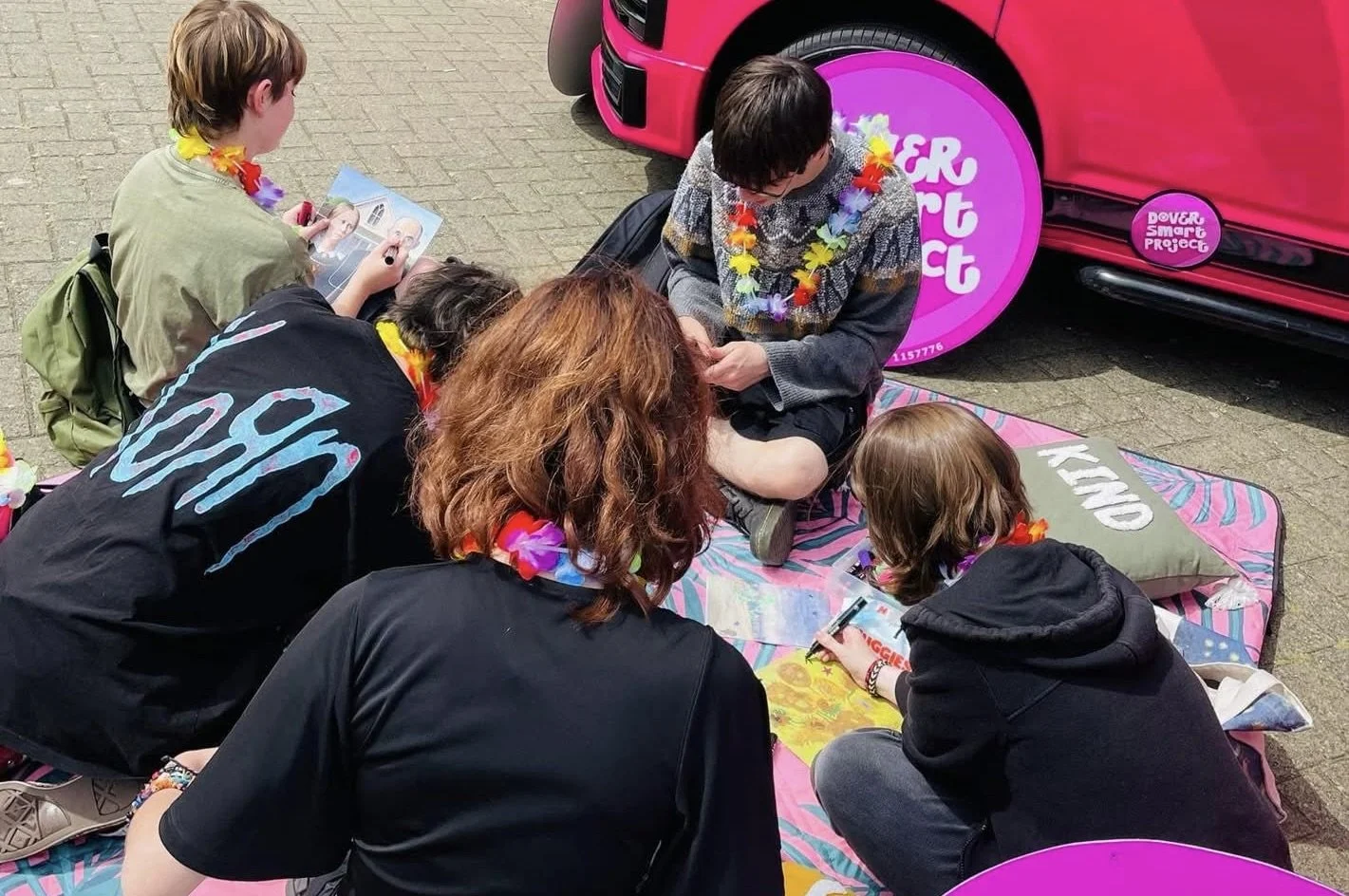 Group of five young people sitting on a colorful blanket on the pavement, wearing leis, engaged in creative activities such as drawing and coloring, with a pink car and a vehicle-themed sidecar in the background, participating in a festive outdoor event.