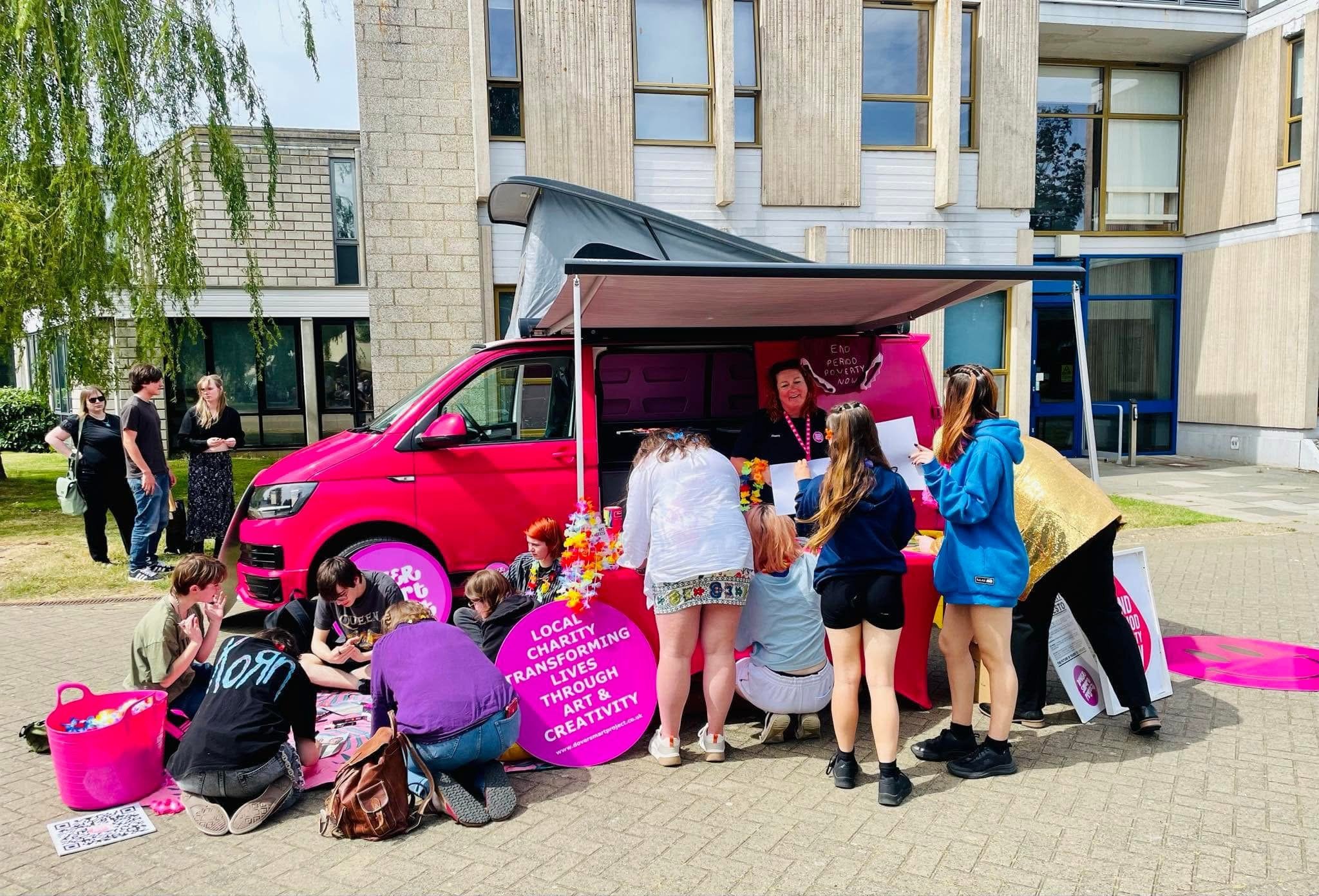 A group of people gathered around a bright pink charity van with a large awning. The van has pink signs with the text 'Local charity transforming lives through art and creativity.' Some individuals are sitting on the ground, while others are standing and interacting. The scene is set outdoors in front of modern apartment buildings with large windows and trees.