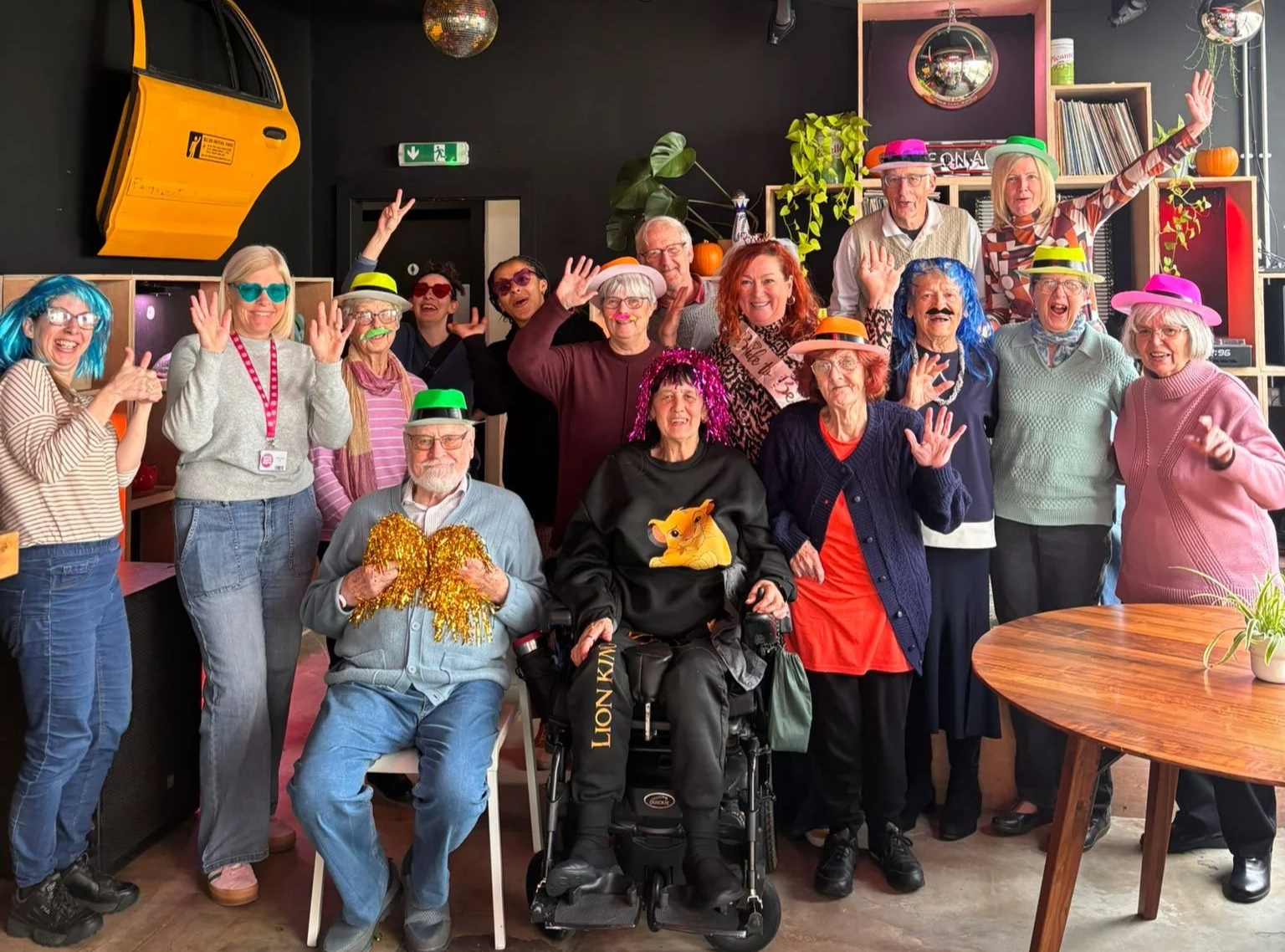 Group of elderly people and a caregiver celebrating together in a decorated indoor space, wearing colorful hats, wigs, and accessories, some waving and smiling, with decorations like pumpkins and plants around.
