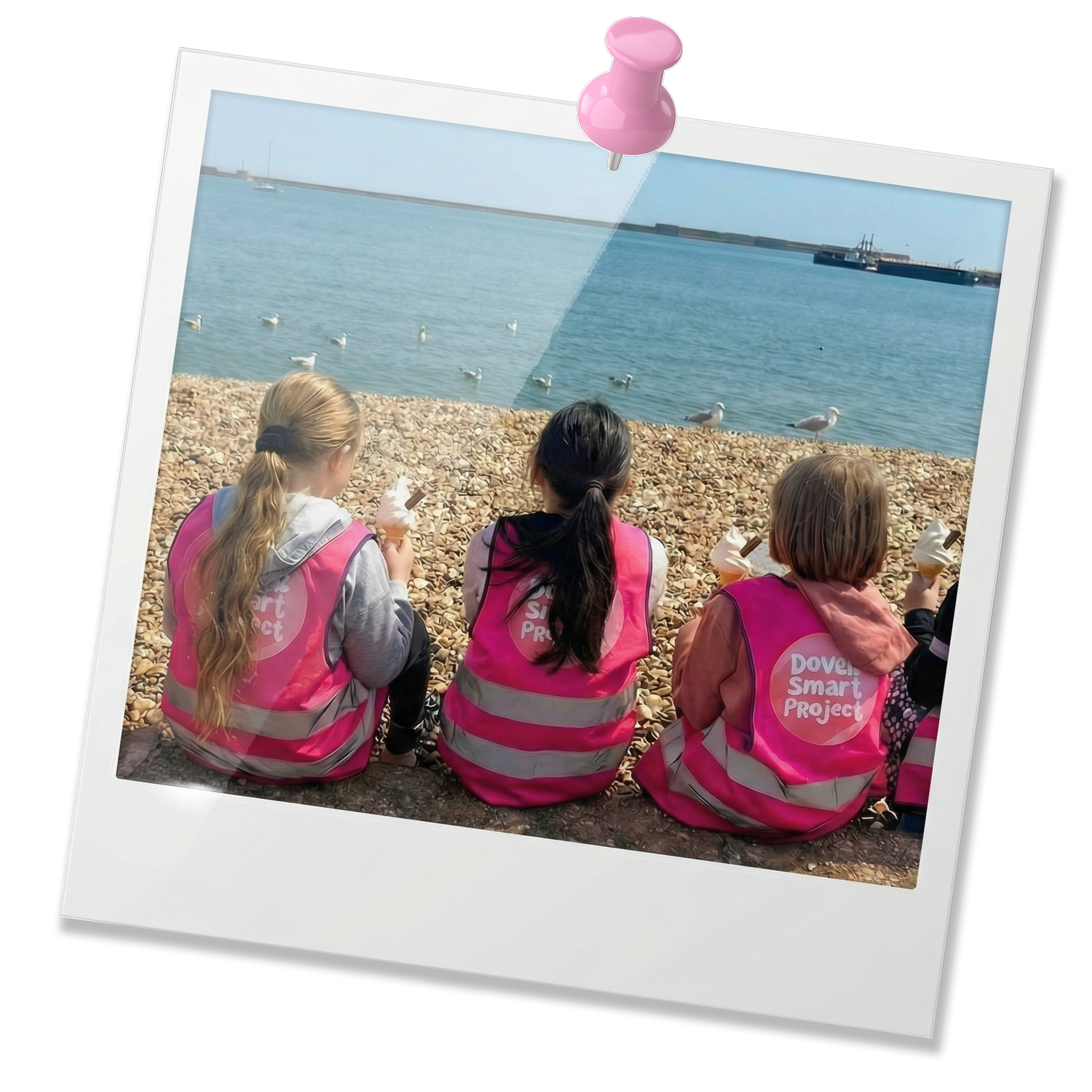 Photo of children sitting on a rocky beach with seagulls and a harbor in the background.