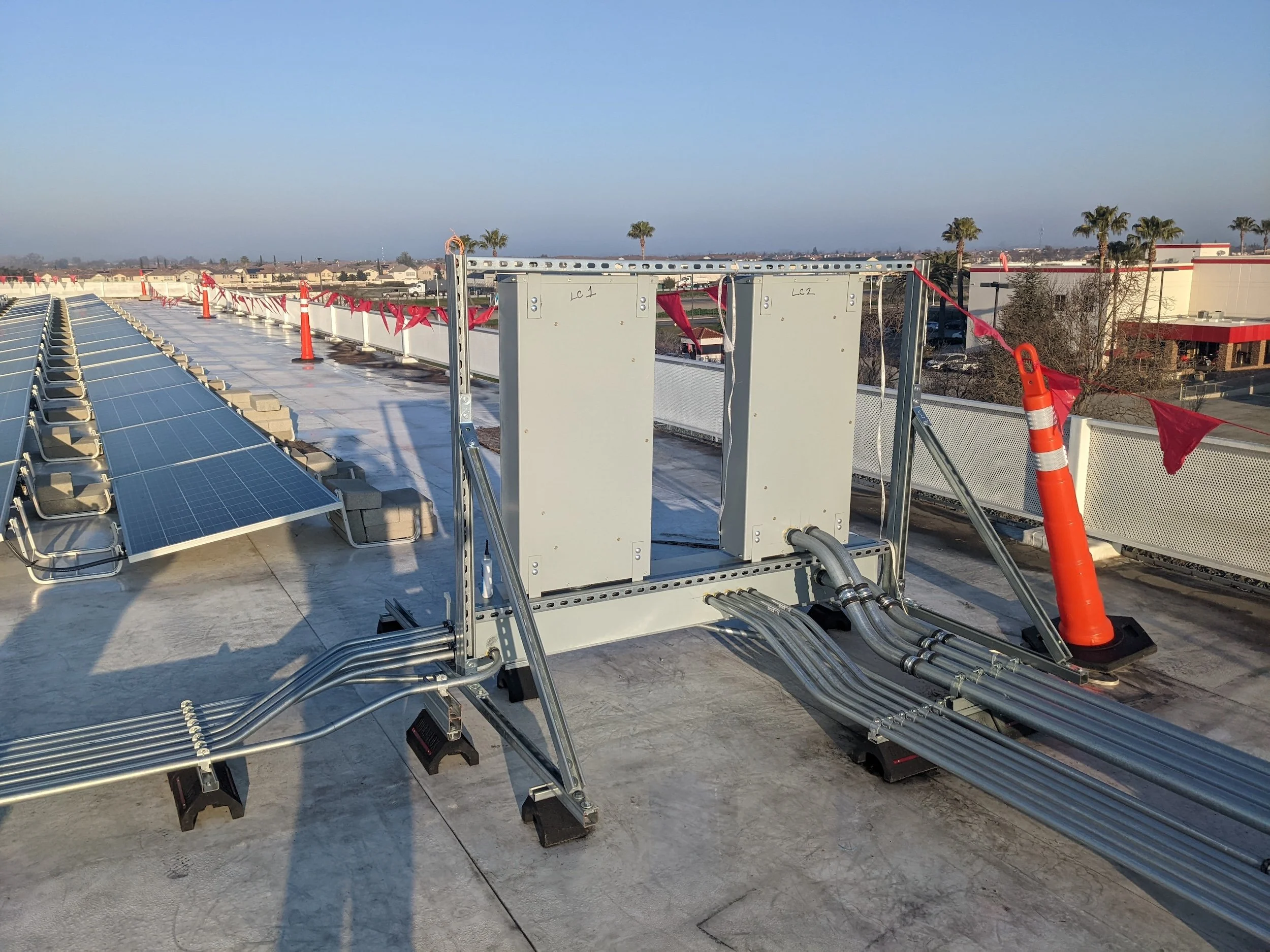 Photovoltaic solar panels and electrical conduits on a rooftop under a clear sky with a cityscape in the background.