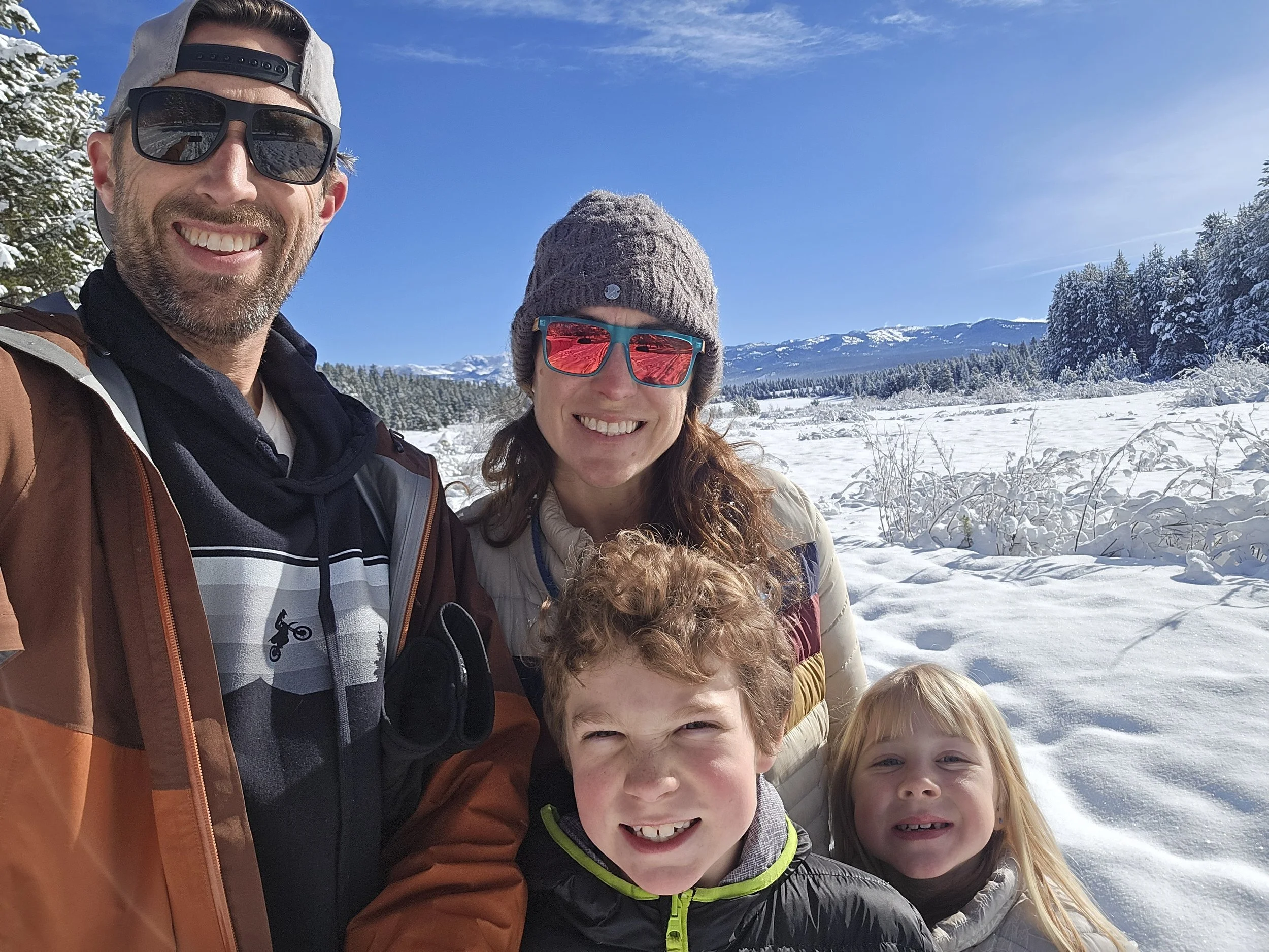 A family of four outdoors in a snowy landscape with mountains in the background, taking a selfie together. The father, mother, and two children are smiling, wearing winter clothing and sunglasses.