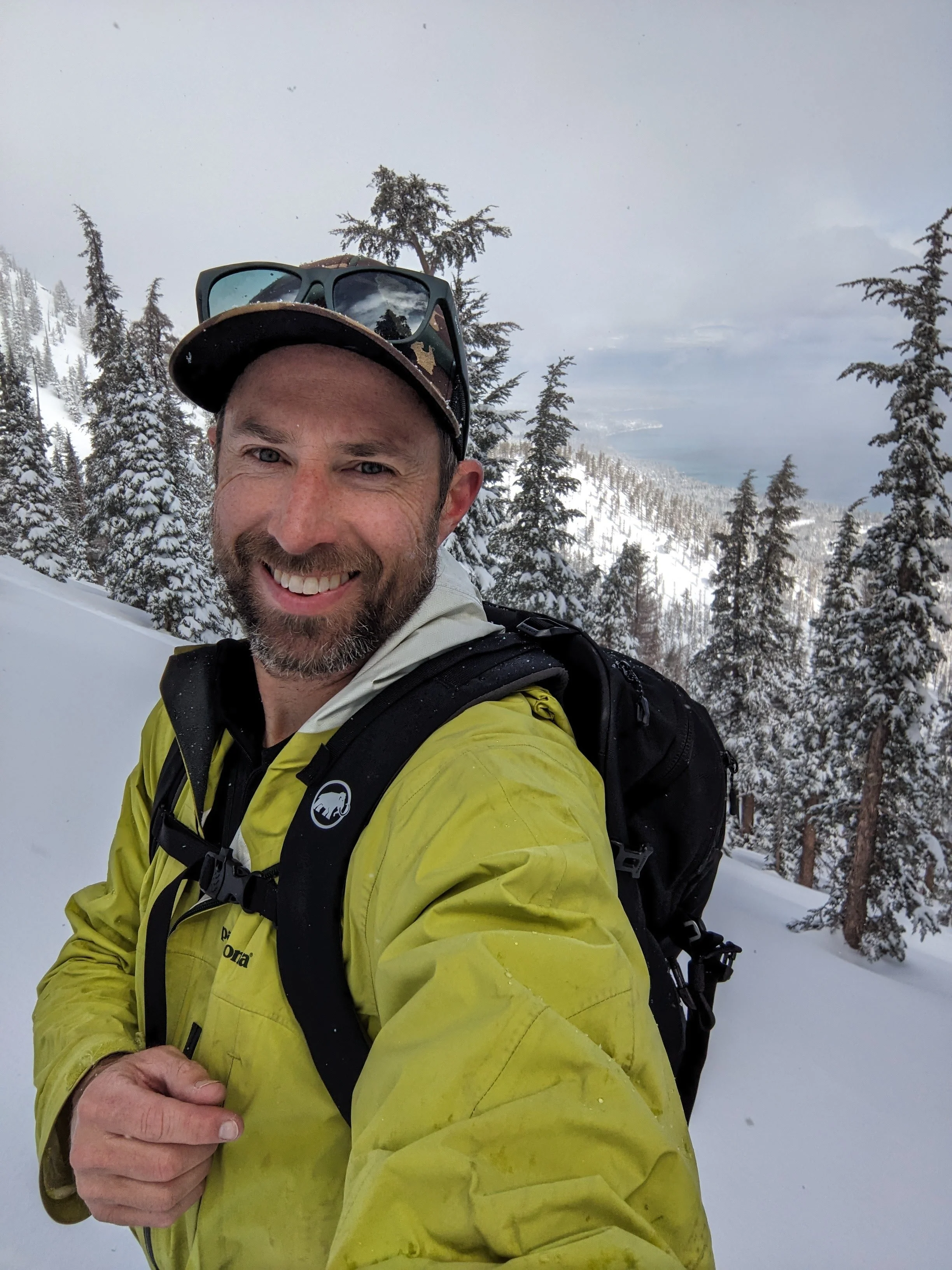 James smiling with a snowy mountain and pine trees in the background, wearing a yellow jacket, black backpack, and sunglasses on his head, taking a selfie in a winter landscape.