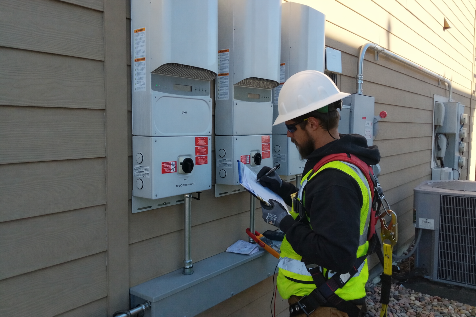 An electrician wearing a safety helmet, sunglasses, gloves, and a high visibility vest is working on electrical equipment mounted on an exterior wall of a building, writing notes on a clipboard.