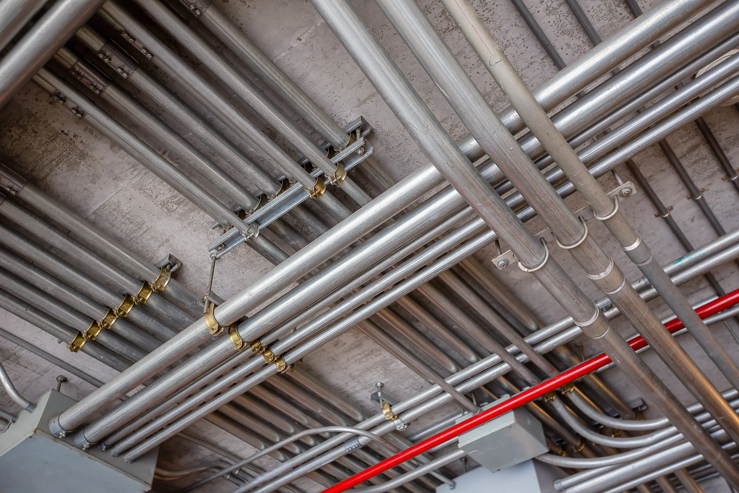 A ceiling with multiple metal pipes and conduits, some secured with brackets and fittings, running parallel and intersecting against a concrete ceiling.