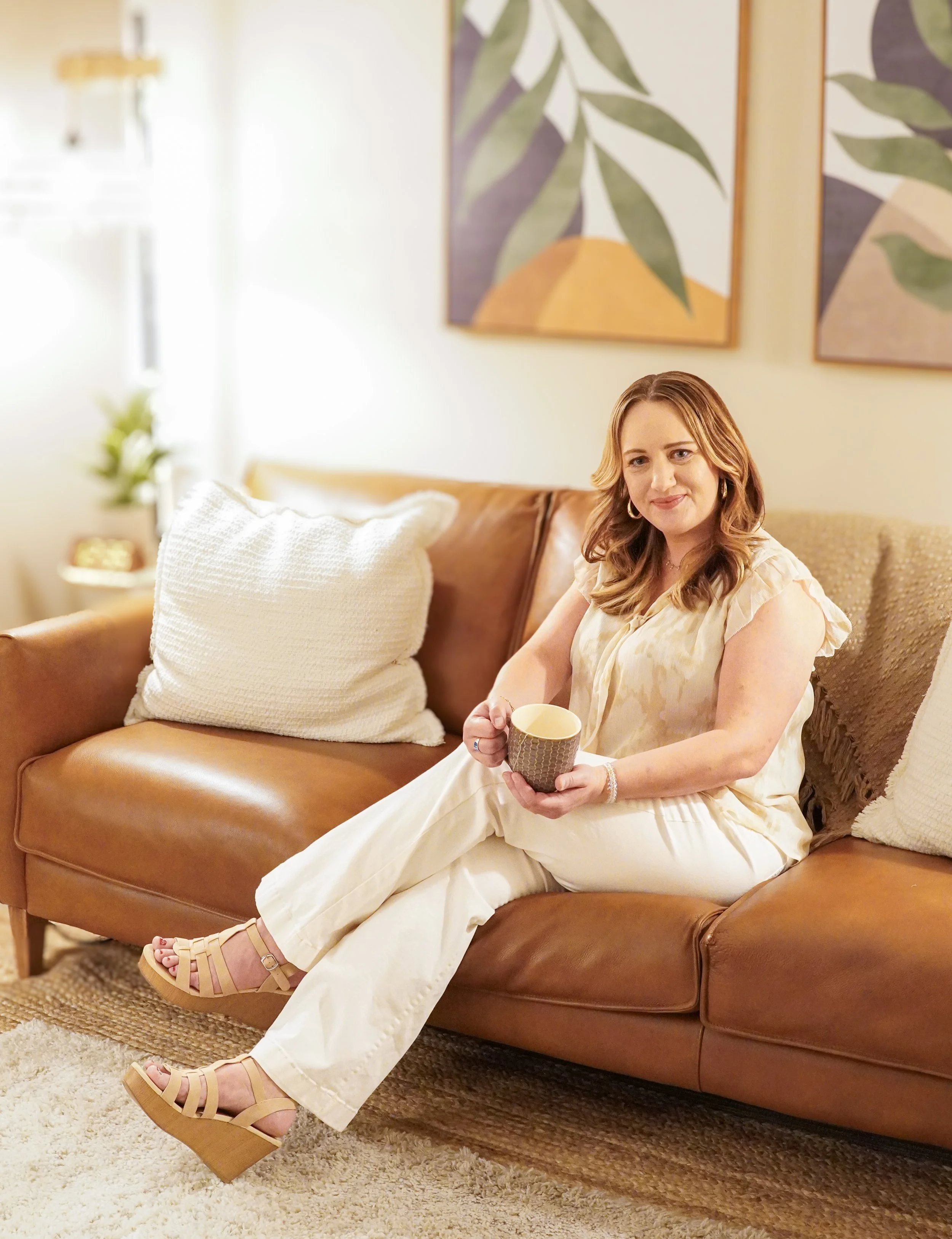 A woman sitting on a brown leather sofa, holding a mug, in a cozy living room with wall art and pillows.