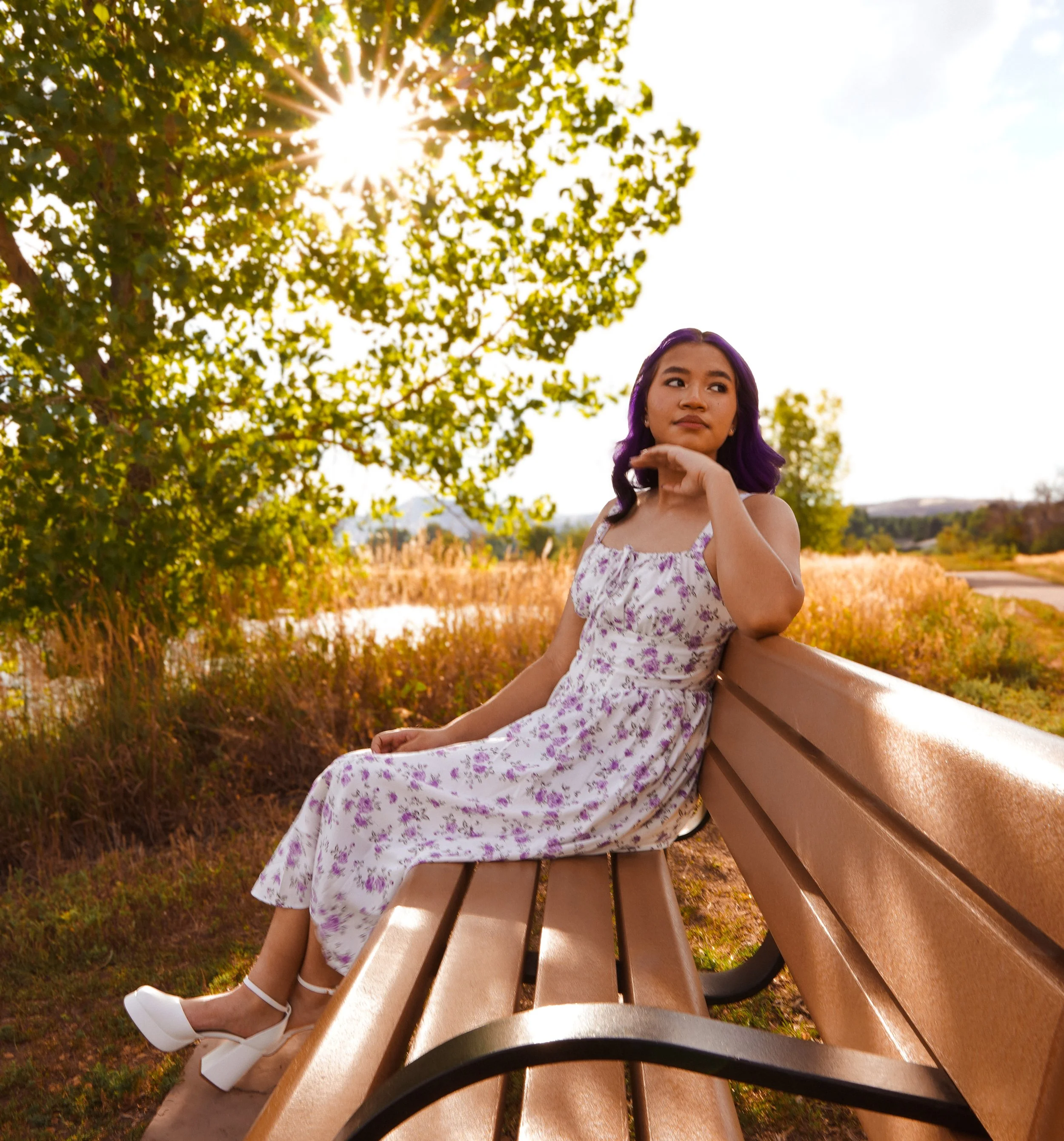 A young woman with purple hair in a floral dress sitting on a park bench, looking off into the distance with the sunlight filtering through the trees in the background.