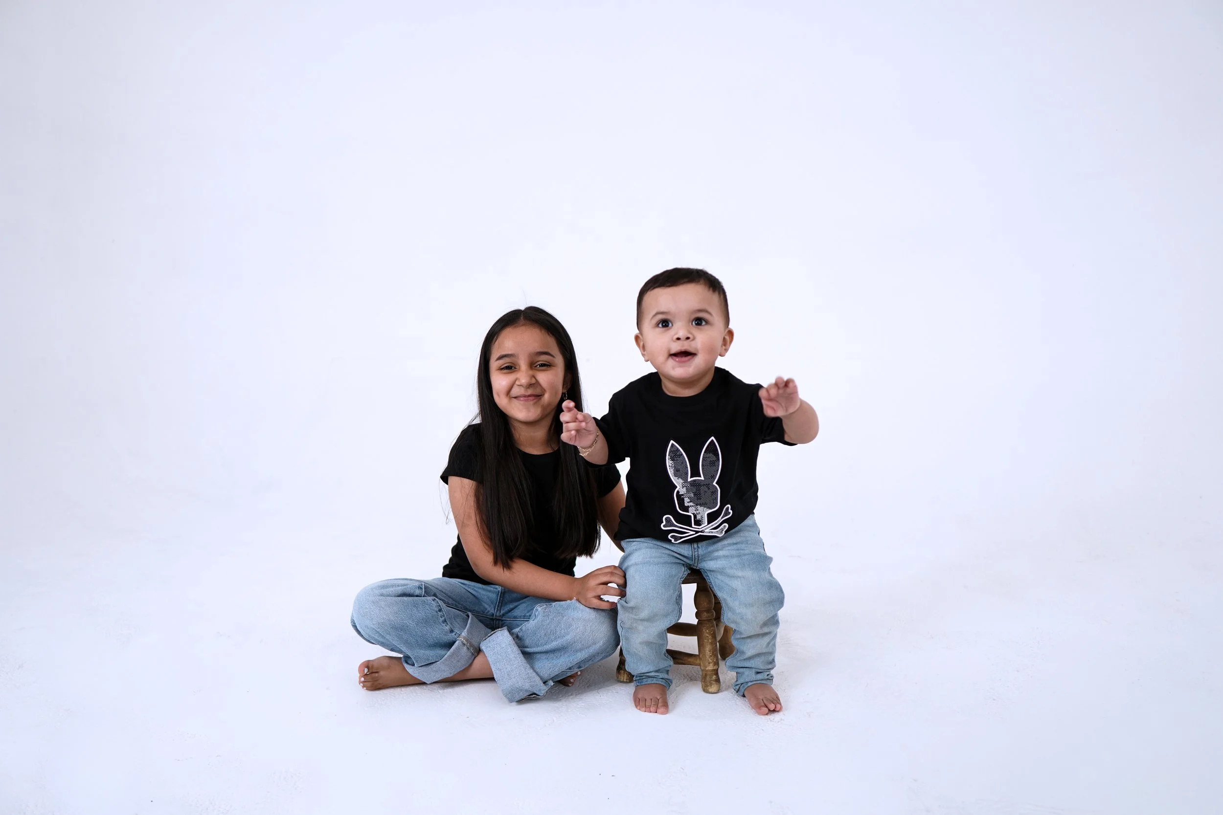A young girl with long dark hair sitting on the floor next to a toddler boy. The boy is sitting on a small wooden stool, wearing a black t-shirt with a bunny logo and light blue jeans, smiling and reaching out. The girl is smiling with her eyes closed, wearing a black t-shirt and jeans. Both are barefoot against a plain white background.