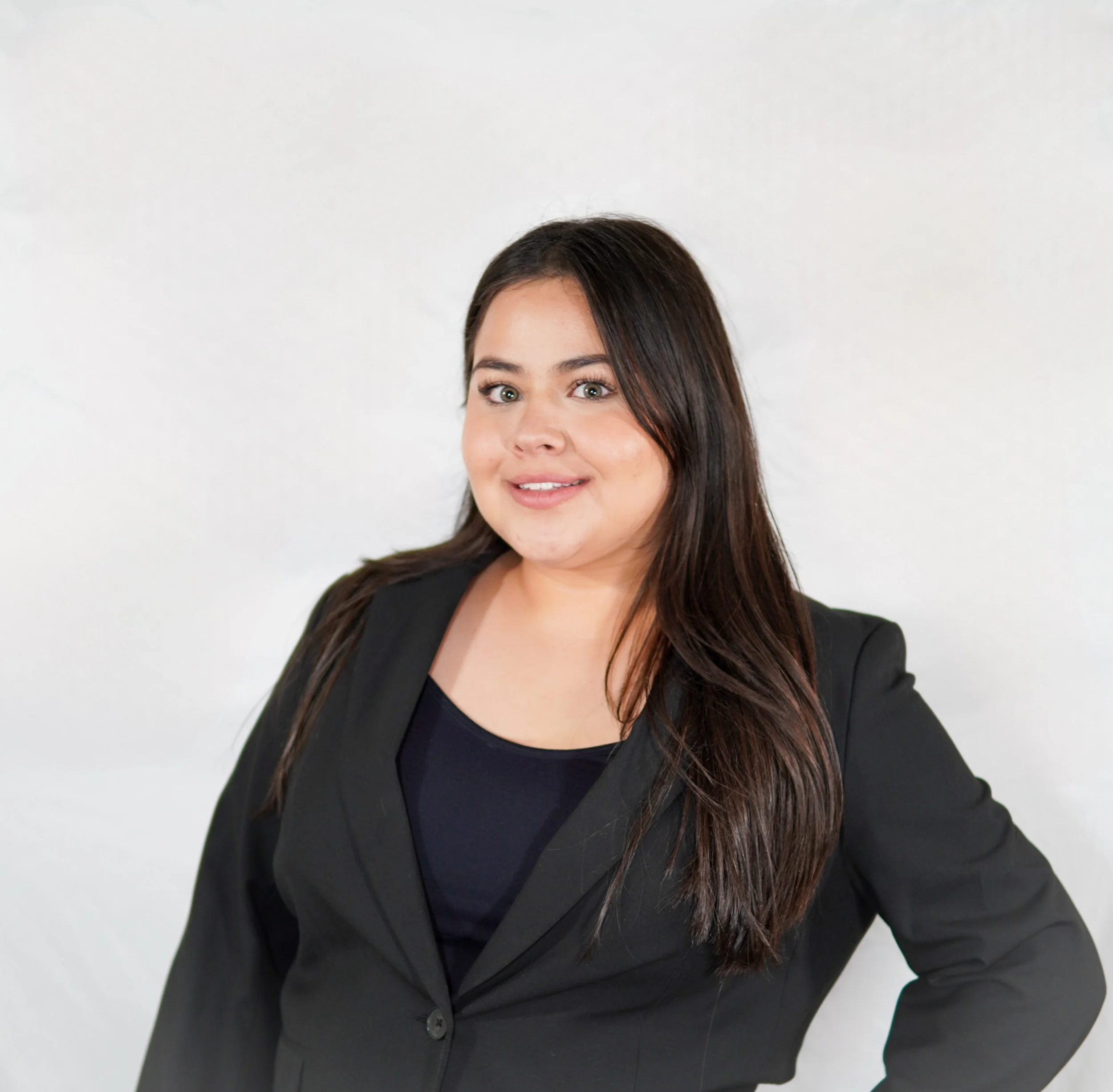Portrait of a woman with long dark hair wearing a black blazer and black top, standing against a plain white background.