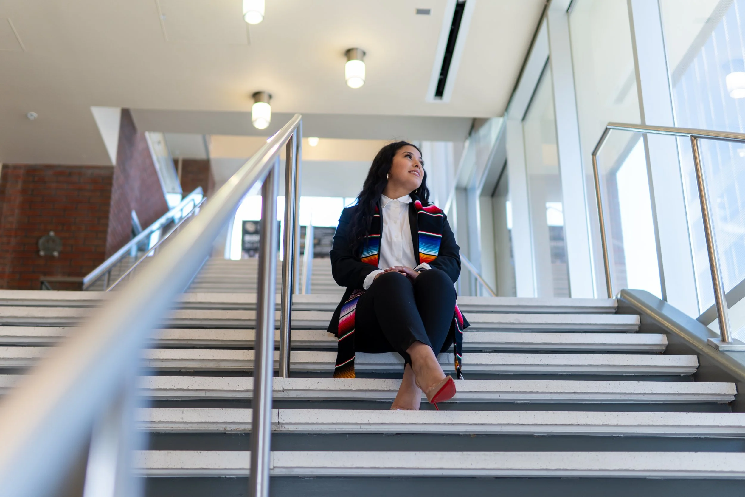 Woman sitting on indoor staircase, wearing a colorful scarf and red high heels, looking out the window.