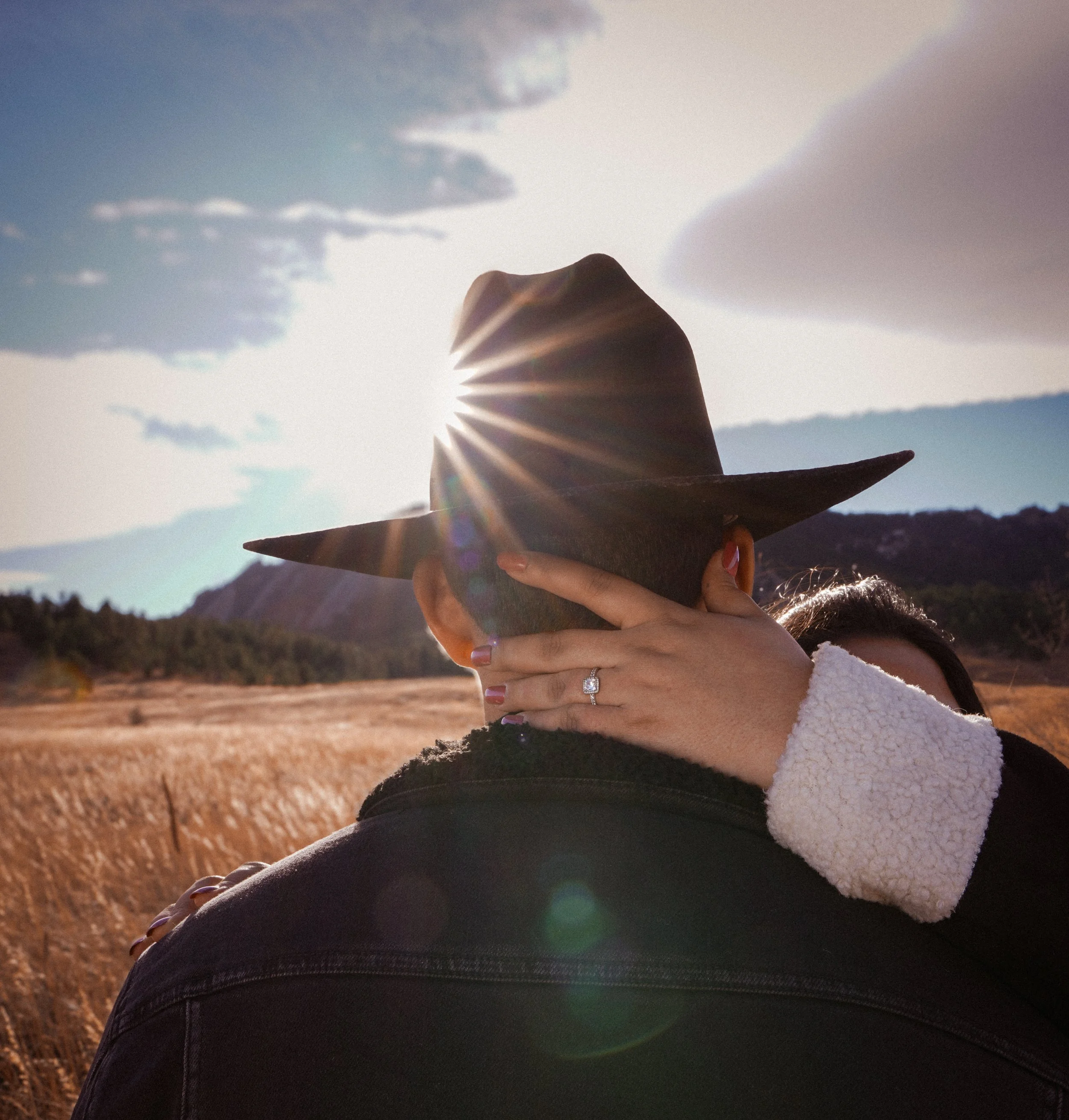 A person wearing a black wide-brimmed hat and a black jacket is embracing someone, with one hand covering the person's face. The person has a ring on their finger. The background shows a field of tall, golden grass with mountains and a partly cloudy sky. The sun is shining brightly, creating lens flare and backlighting the scene.