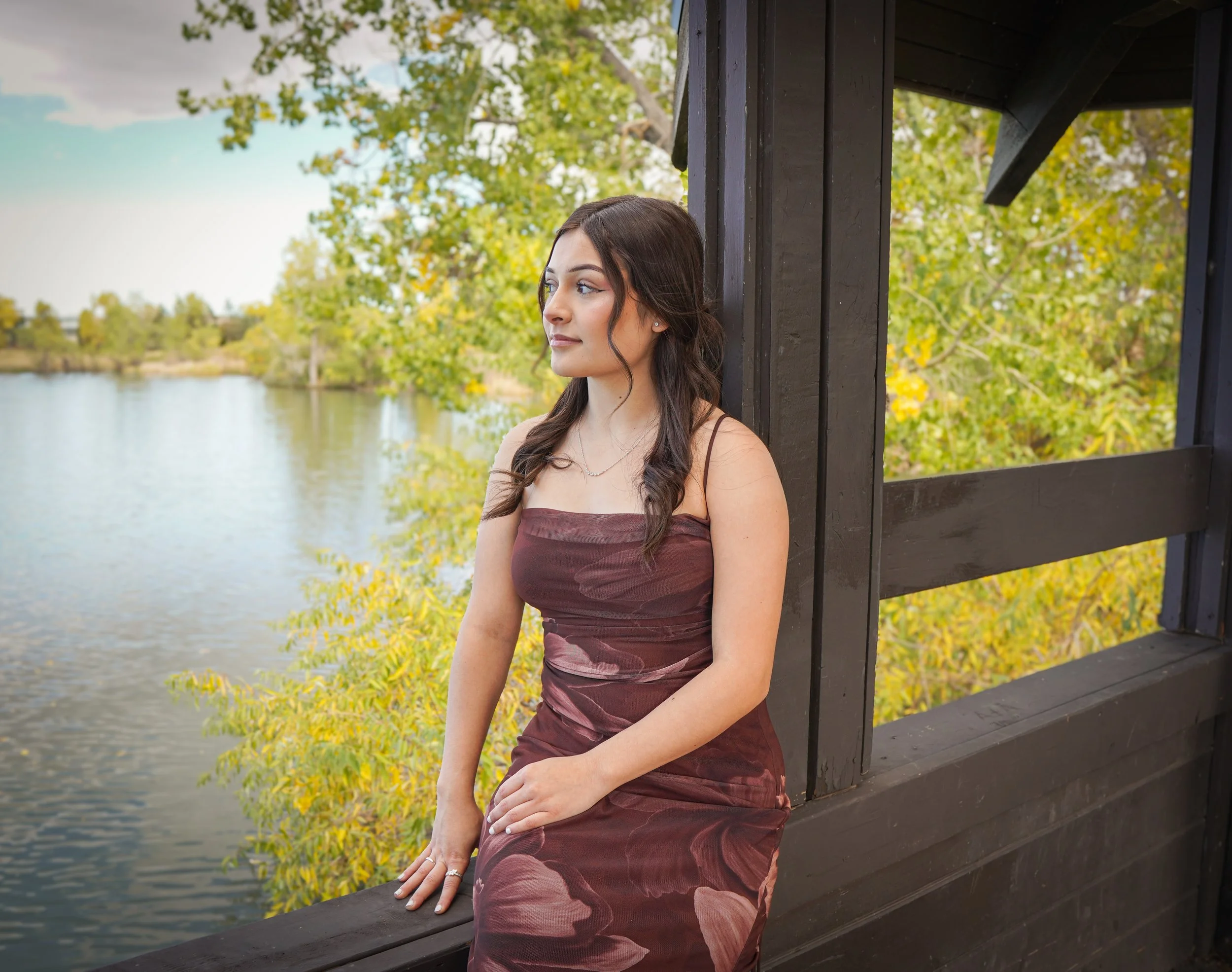 A woman with long dark hair in a strapless dress sitting on a wooden railing near a body of water, with trees and leaves in the background.