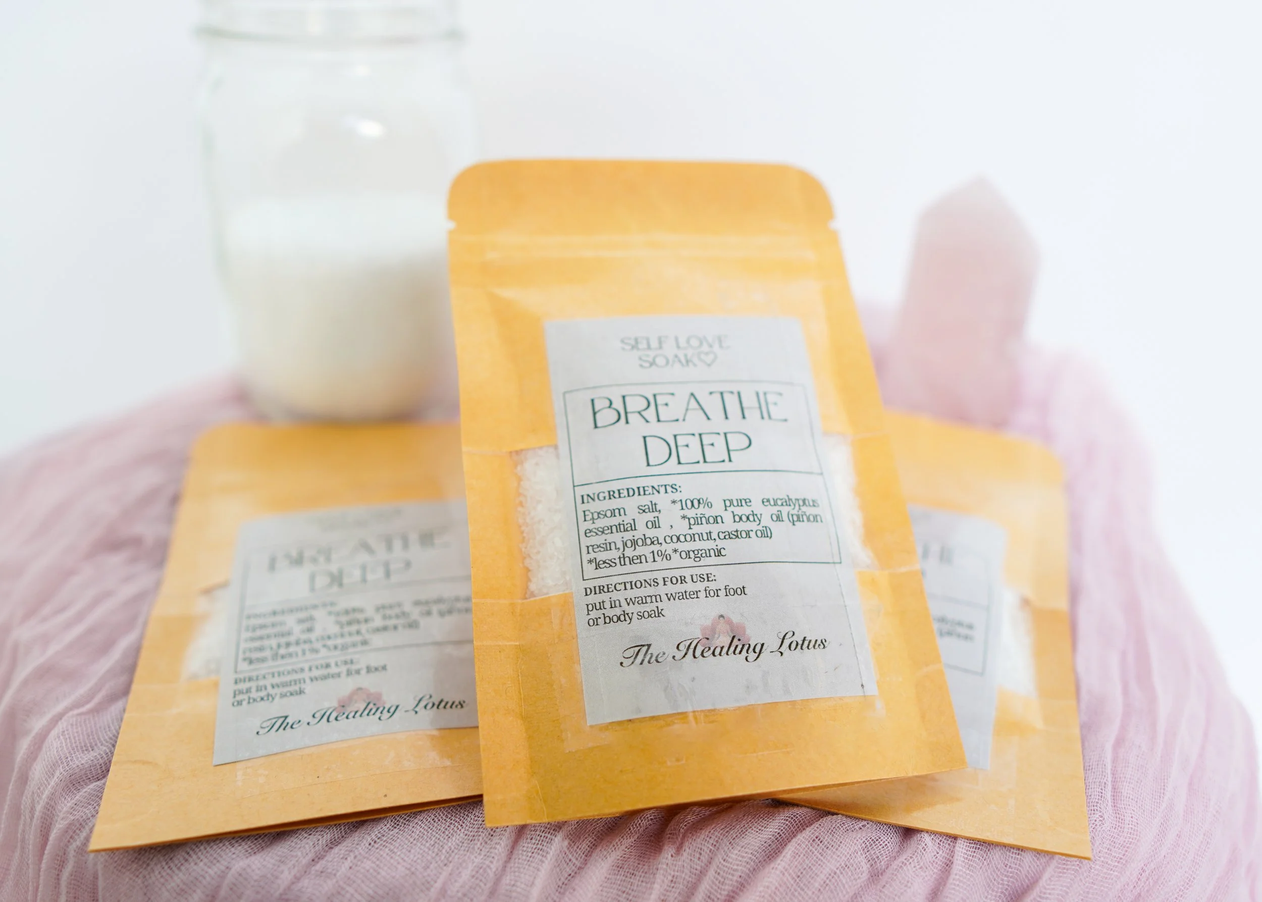 Three orange pouches labeled 'Breathe Deep' with ingredients and usage instructions, one clear jar of white bath salts or sugar, and pink bath stones, all on a pink textured cloth.