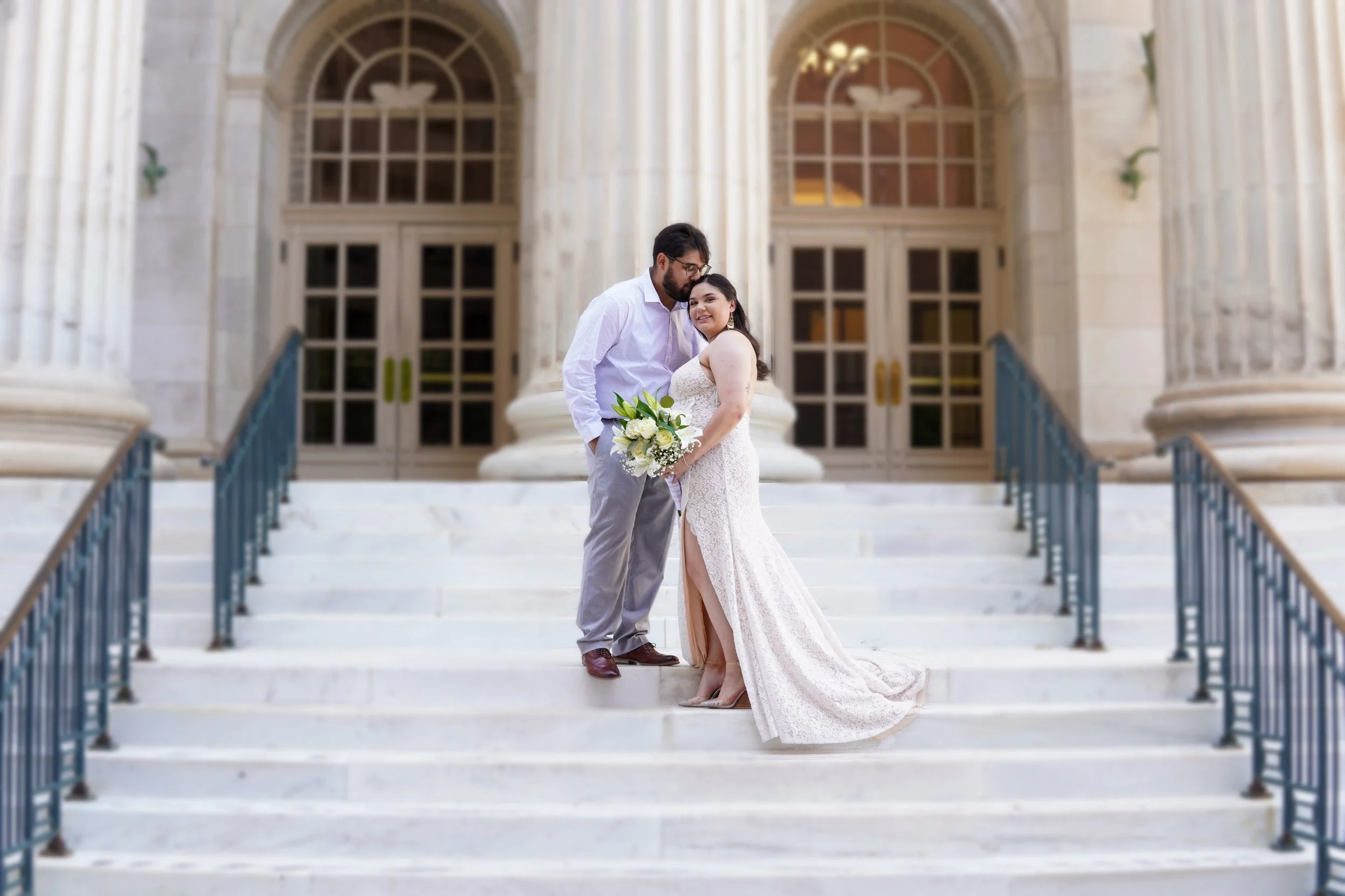 A couple in wedding attire holding a bouquet on marble stairs outside a classical building with columns and arched windows.