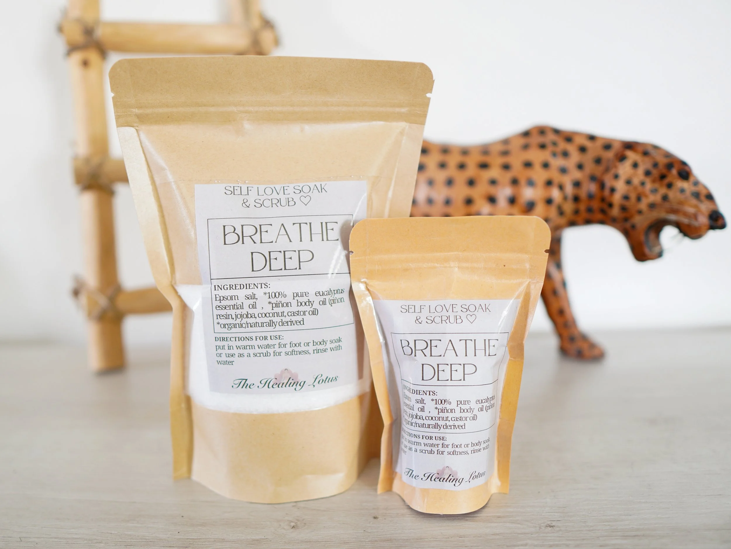 Two beige resealable pouches labeled "Breathe Deep" self-love soak and scrub, with a decorative giraffe figurine in the background.