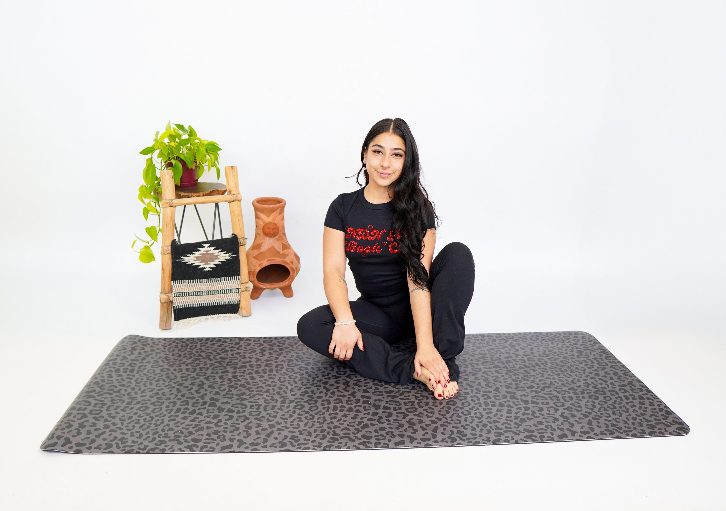 A woman with long black hair, wearing a black T-shirt with red lettering and black pants, sitting cross-legged on a gray patterned yoga mat in a white studio, with a small wooden shelf holding a potted plant and a decorative terracotta piece in the background.