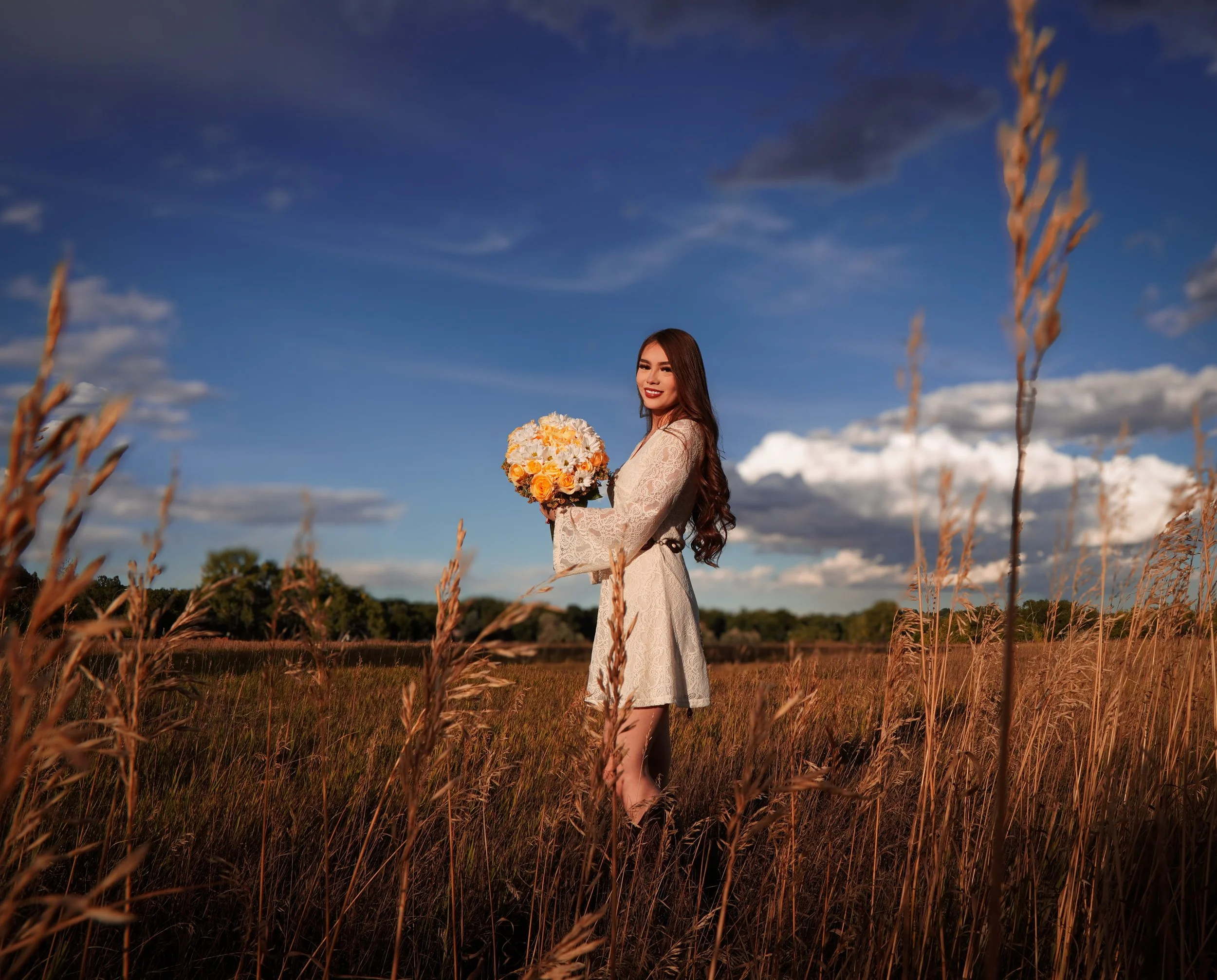 A woman in a white dress holding a bouquet of flowers in a field of tall grass, with a blue sky and white clouds overhead.