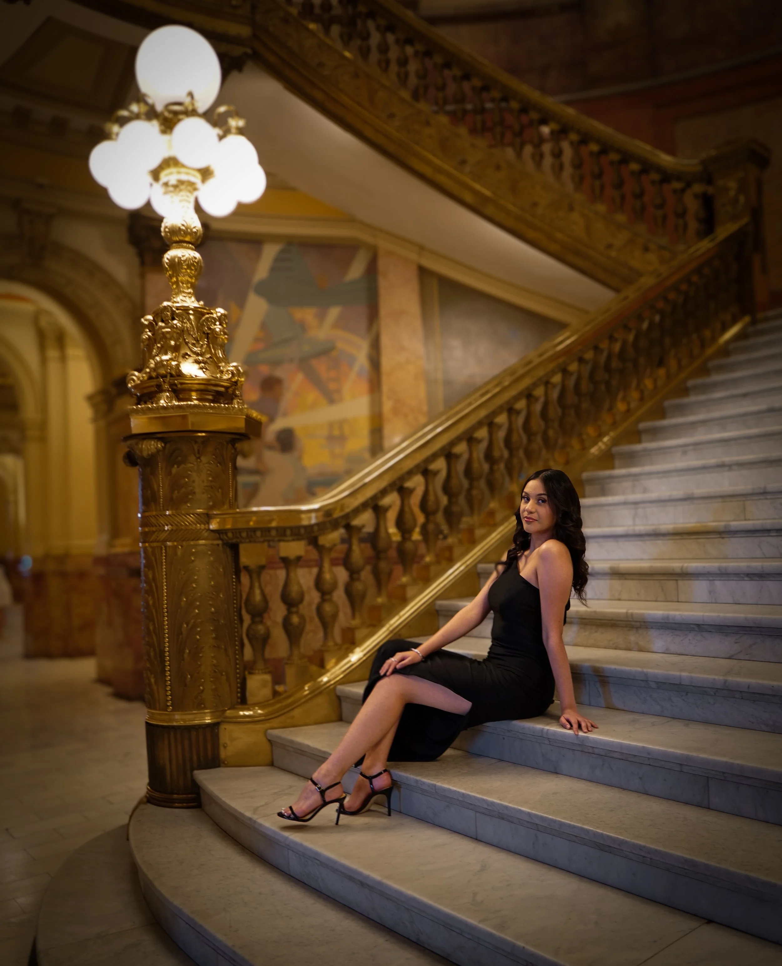 A woman in a black dress sitting on marble stairs inside a grand, ornate building with gold accents and an elaborate staircase banister.