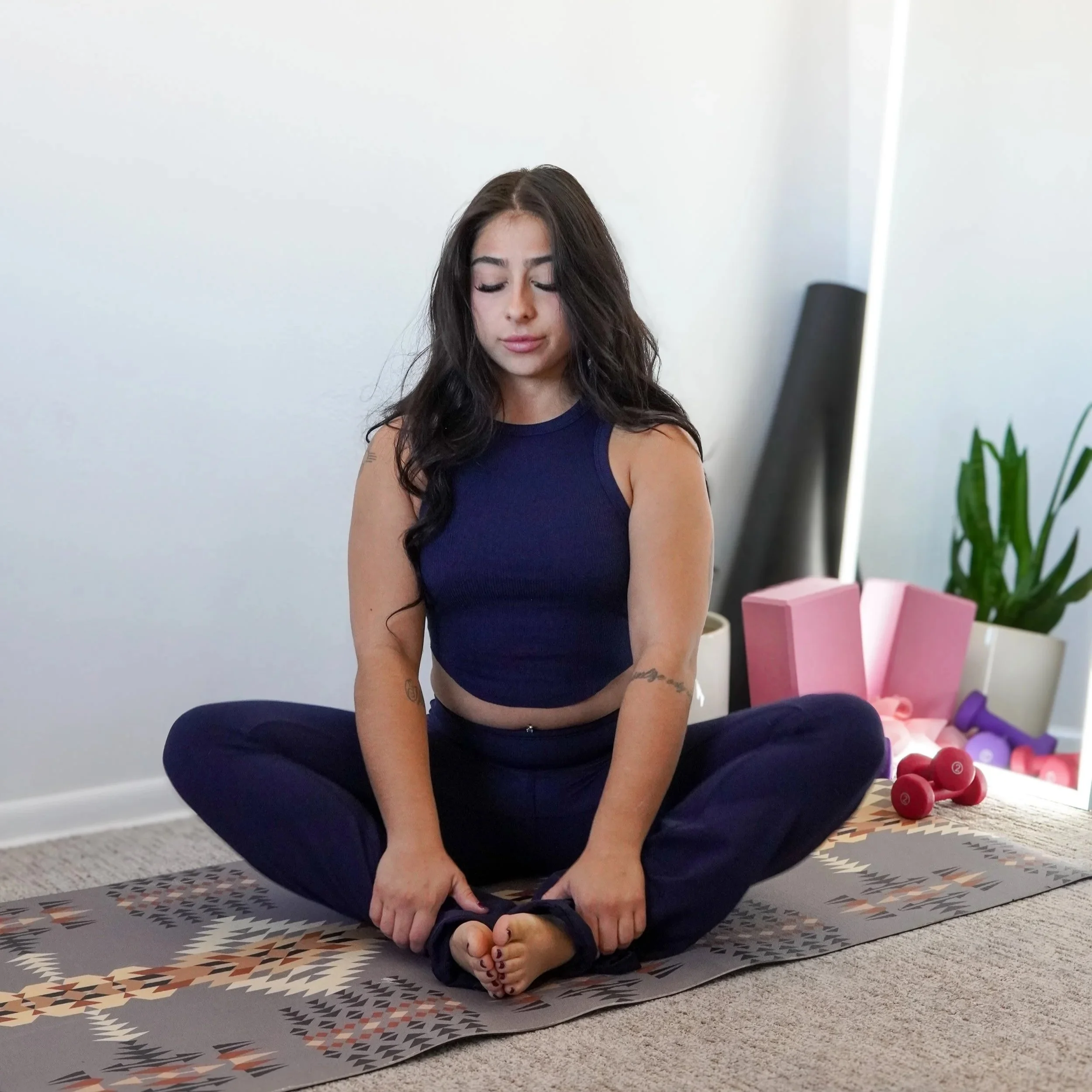 A young woman with long dark hair in a blue sports bra and navy yoga pants sitting cross-legged on a patterned yoga mat, holding her feet with a calm expression, in a room with a white wall, a plant, pink gift boxes, and colorful dumbbells.