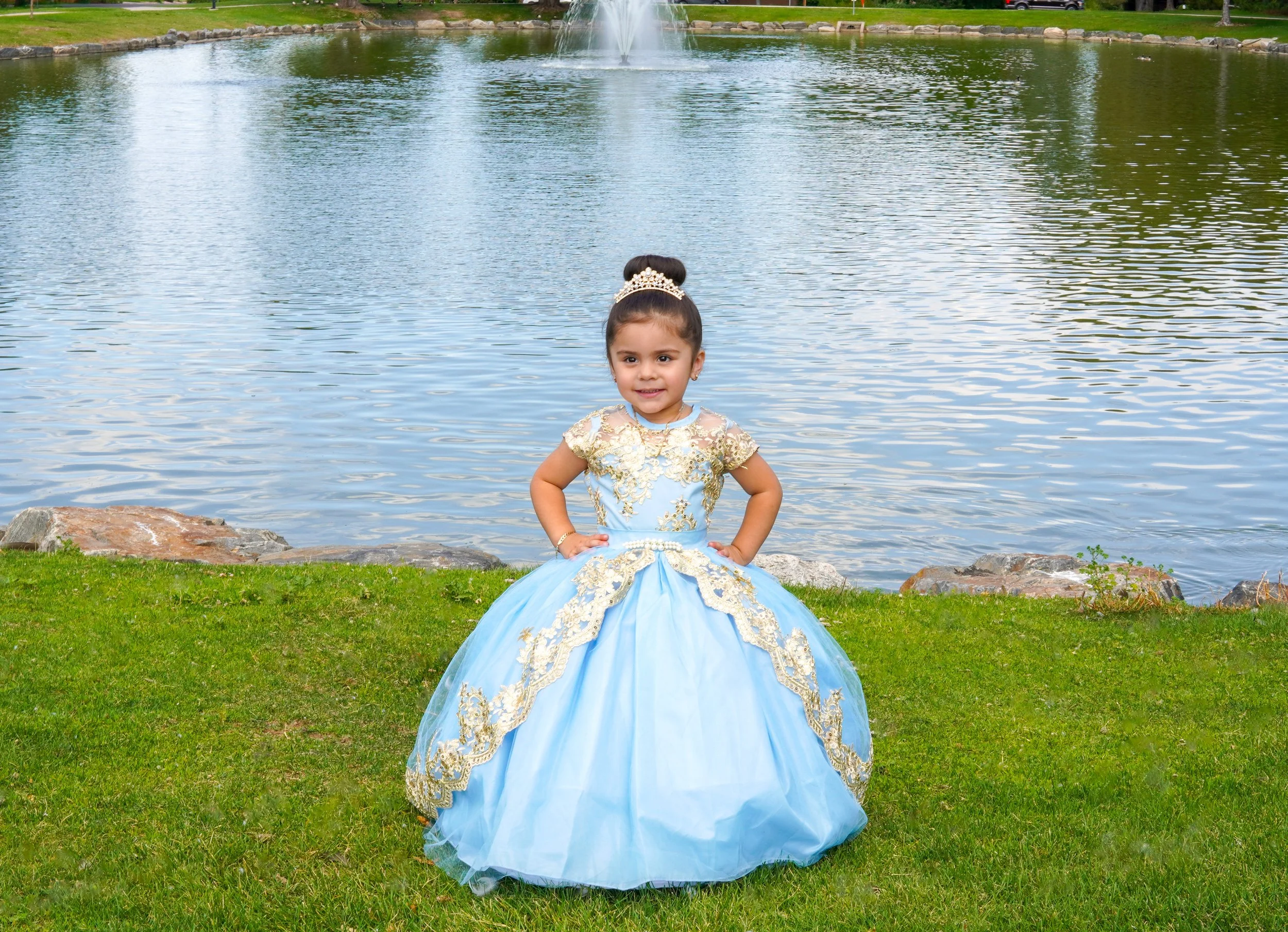 Young girl dressed as a princess in a light blue gown with gold embroidery, standing on grass near a pond with rocks and a fountain in the background.