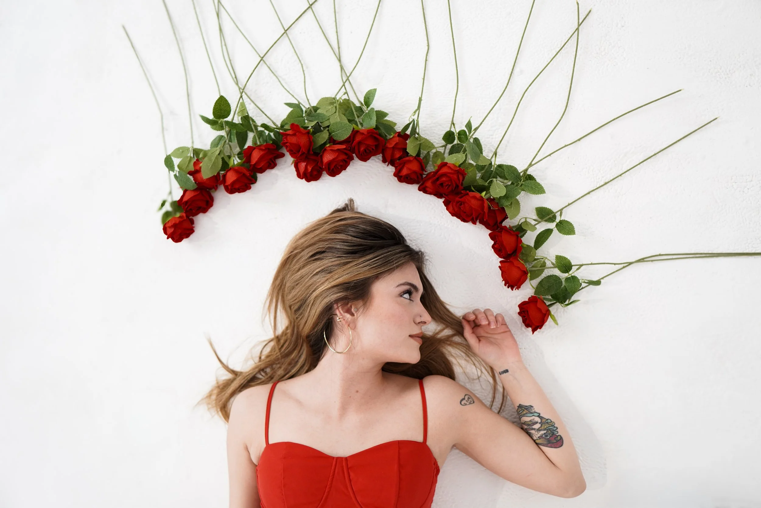 A woman with long, light brown hair and tattoos on her arm wearing a red spaghetti strap dress, lying on a white wall with a semi-circle arrangement of red roses and green leaves above her head.