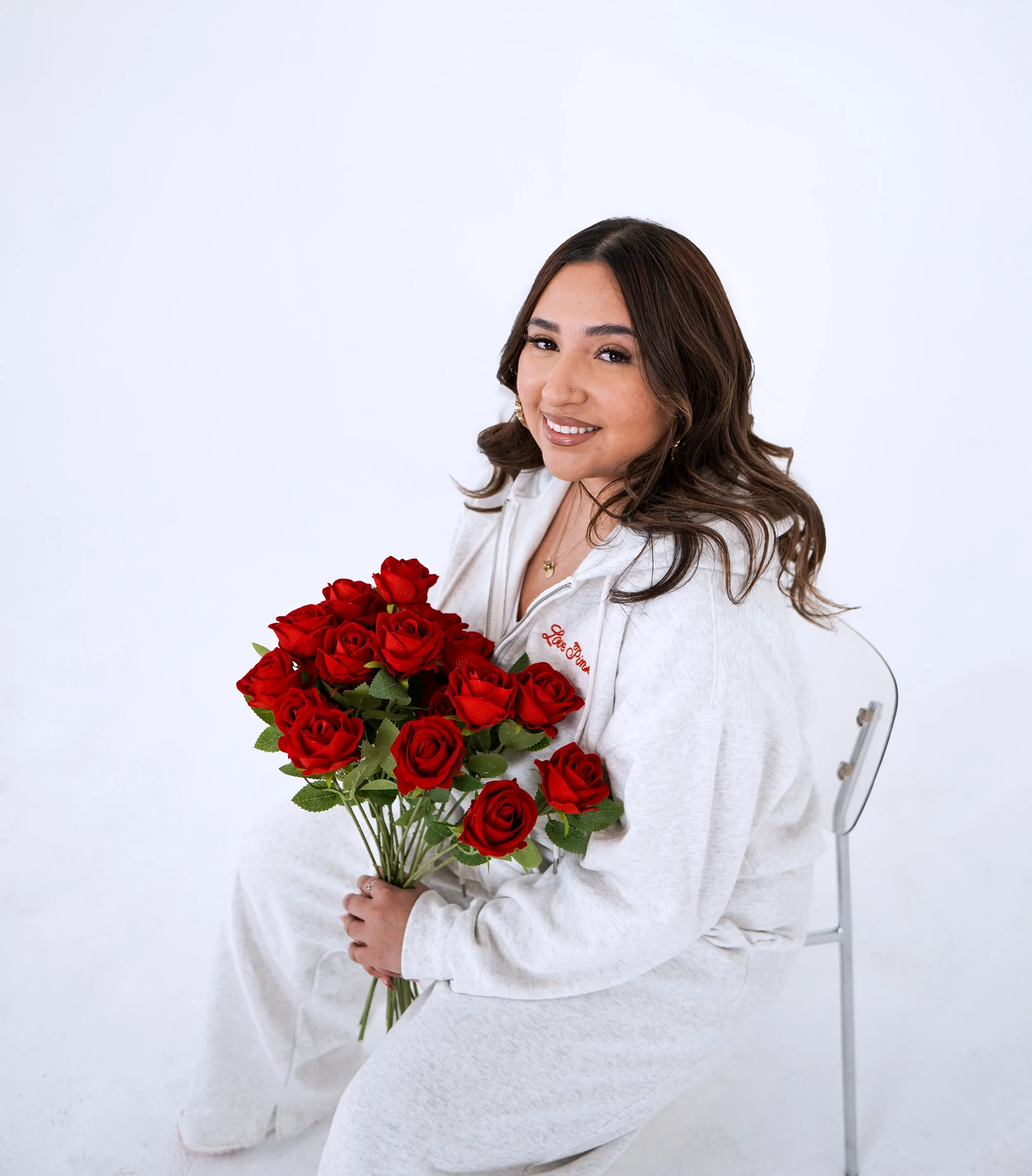 A woman with brown hair and a white outfit sitting on a transparent chair and holding a bouquet of red roses, smiling at the camera.