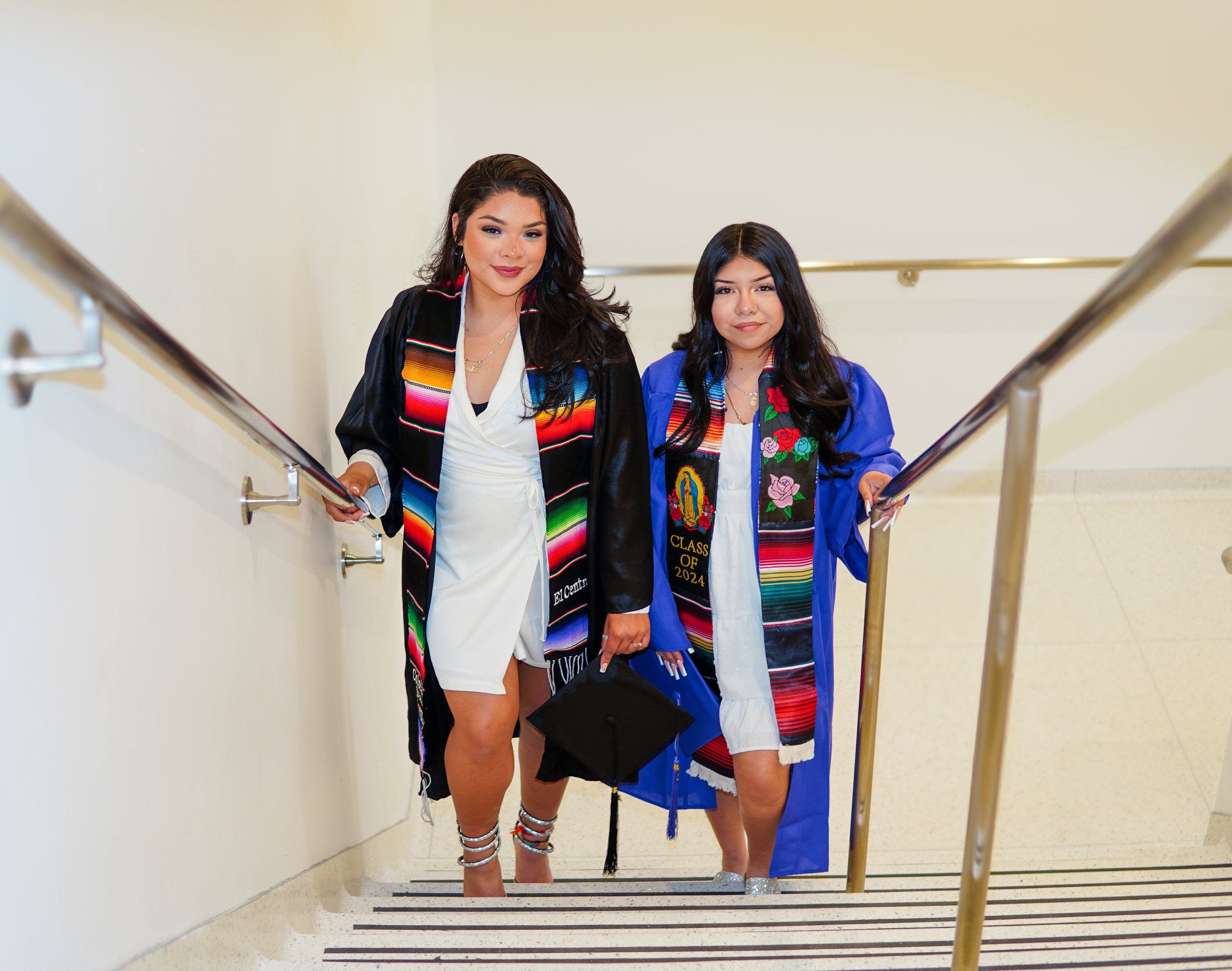 Two young women in graduation gowns and stoles walking up a staircase inside a building, celebrating graduation.