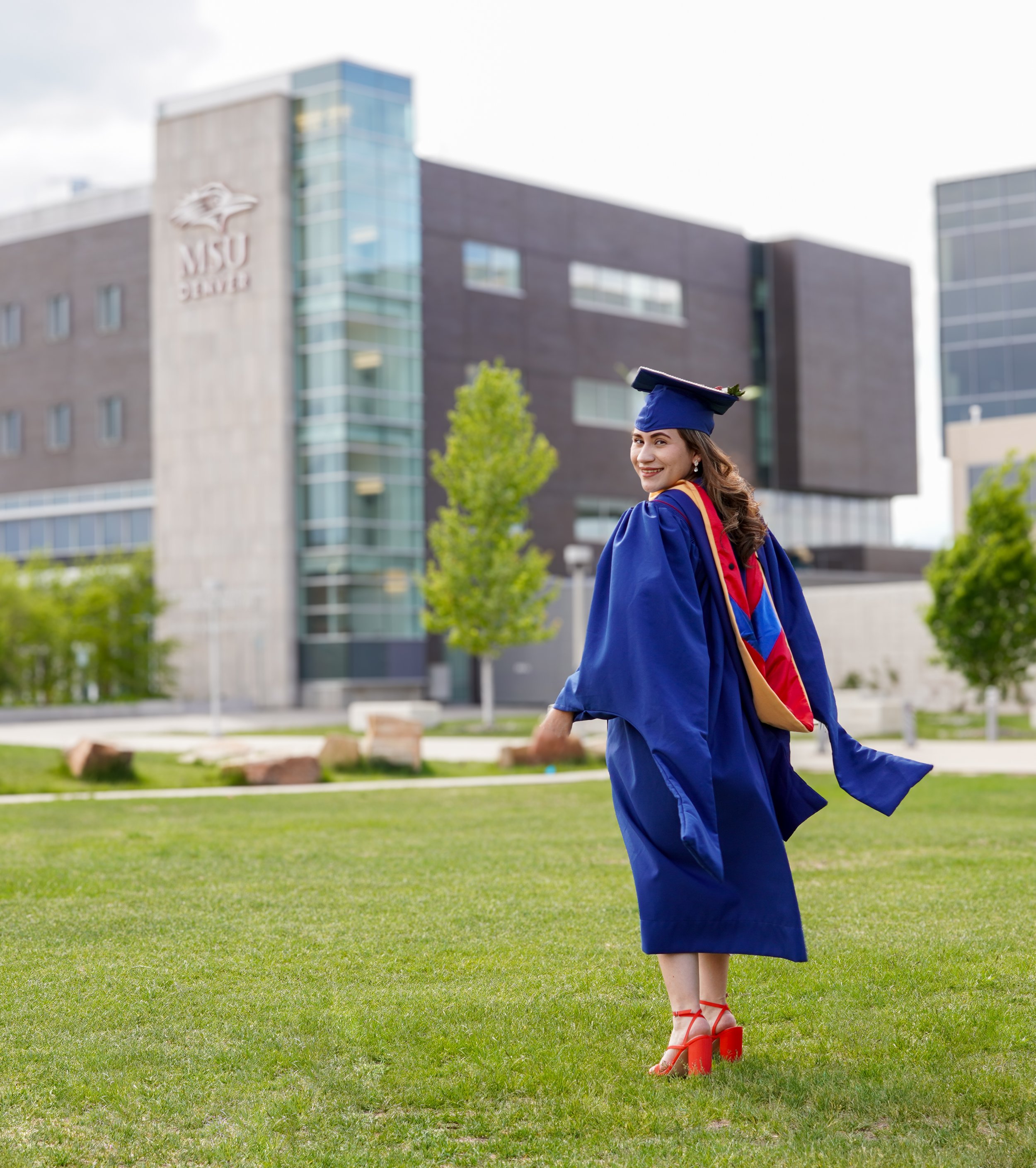 A woman in a blue graduation gown and cap smiling and walking on grass with a university building in the background.