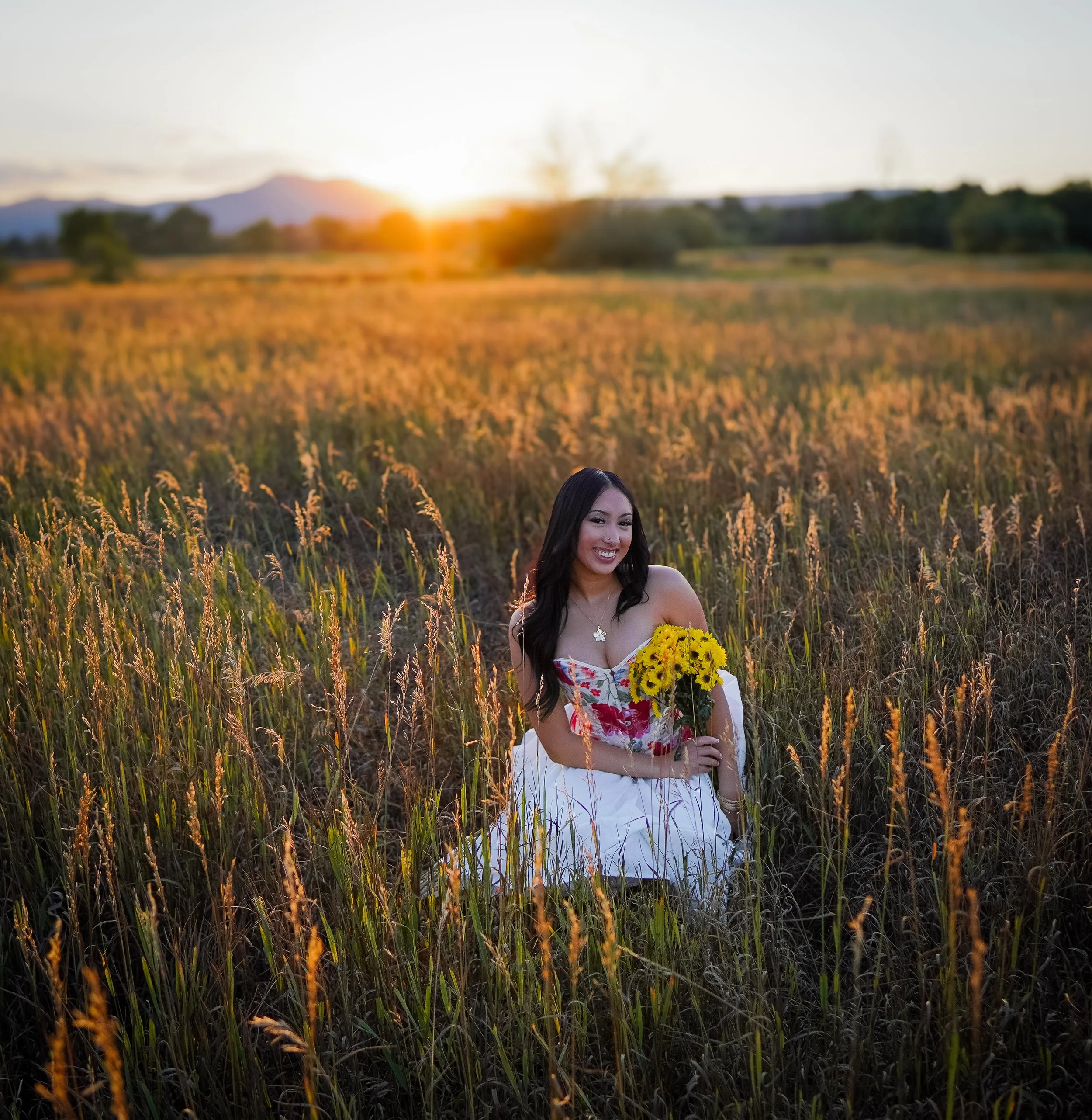 A woman smiling while sitting in a field of tall grass during sunset, holding a bouquet of yellow sunflowers.